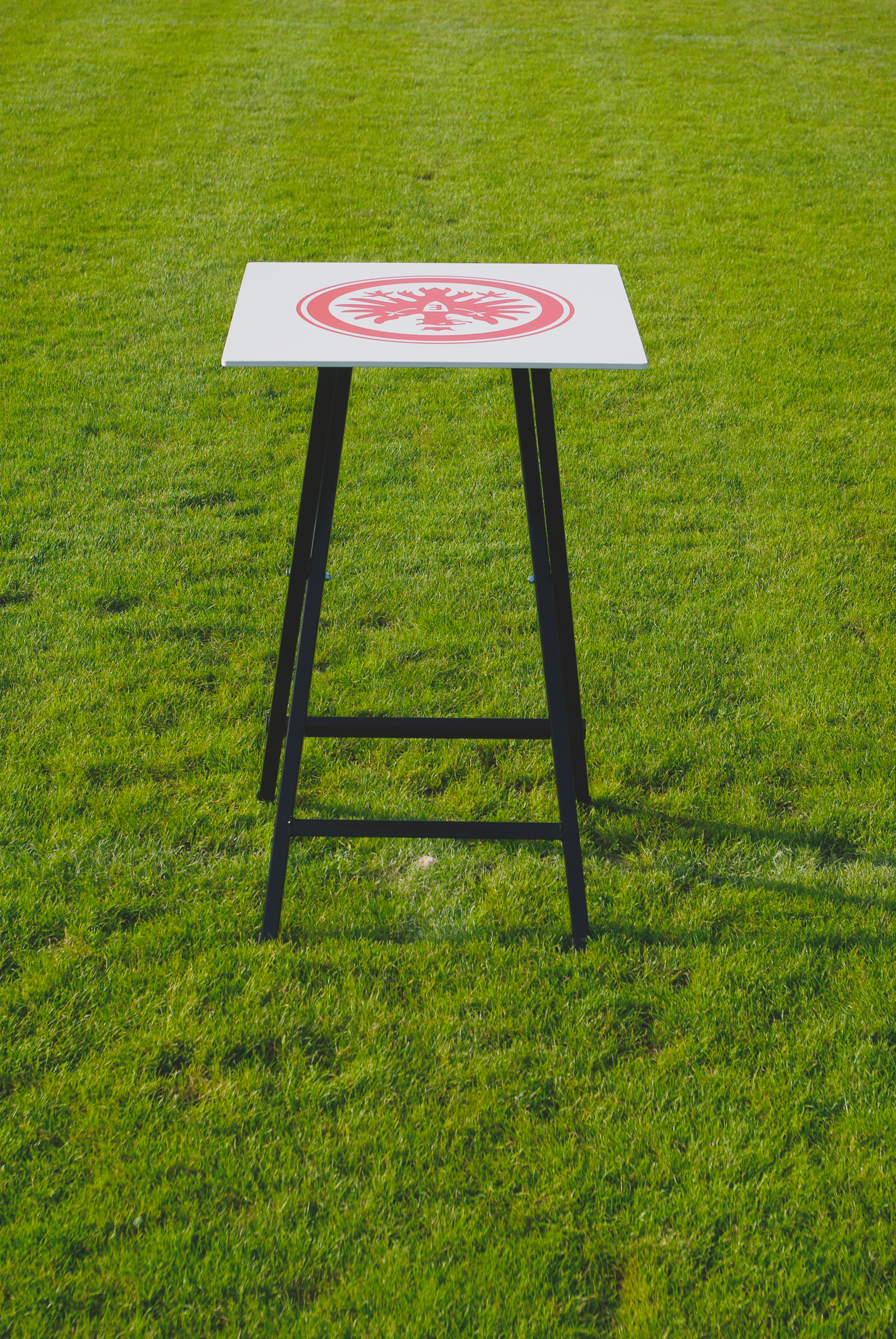 Weißer Stehtisch mit rotem Adler-Emblem auf Sportplatz