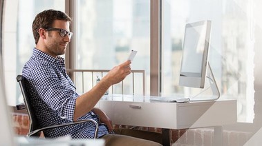 A man seated at a desk, focused on a computer while holding a cell phone in his hand.