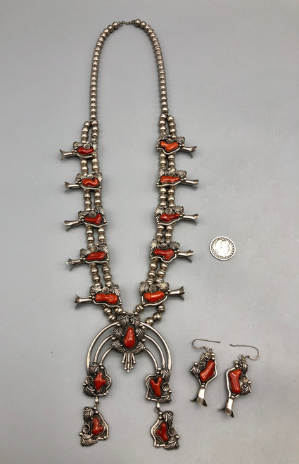 Offered here is a ravishing red coral squash blossom necklace and earrings set. This is an exquisite presentation featuring the 70s style of silver floral applique that wraps beautifully around each branch coral setting. The naja has two beautifully matching dangles and the hook earrings go perfectly with the necklace. It is strung on a fine chain and is stamped sterling. 
MAKER: Hallmarked D.H. (stamped in script).
APPROX AGE: Circa 1970s.
SIZE: Measures 30 inches end to end. Pendant measures 4-1/2 inches long X 2-7/8 inches wide. Earrings measure 1-7/8 inches long X 7/8 inches wide. Weighs 212 grams. 
CONDITION: Good for its age, normal wear expected. Please look closely at the pictures as they are an important part of our description and are hereby incorporated into the description, specifically with regards to condition.