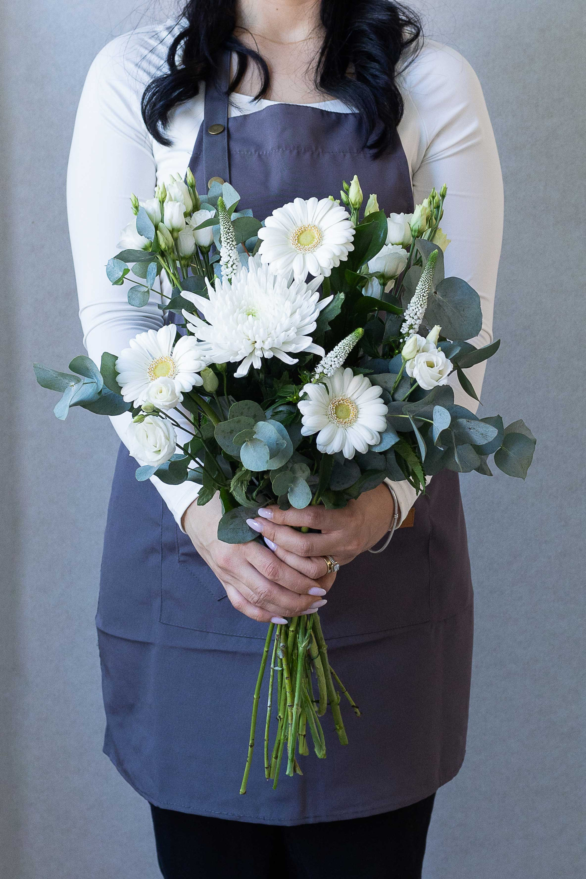 Florist holding a bouquet of white gerbera, chrysanthemum, lisianthus, veronica, and eucalyptus foliage against a gray background.