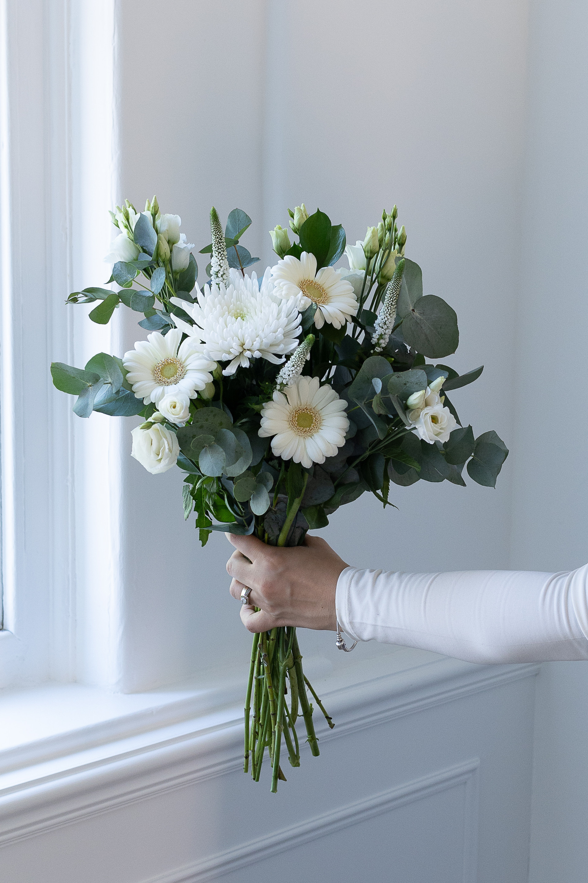 Hand holding a bouquet of white gerbera, chrysanthemums, lisianthus, and eucalyptus beside a bright window.
