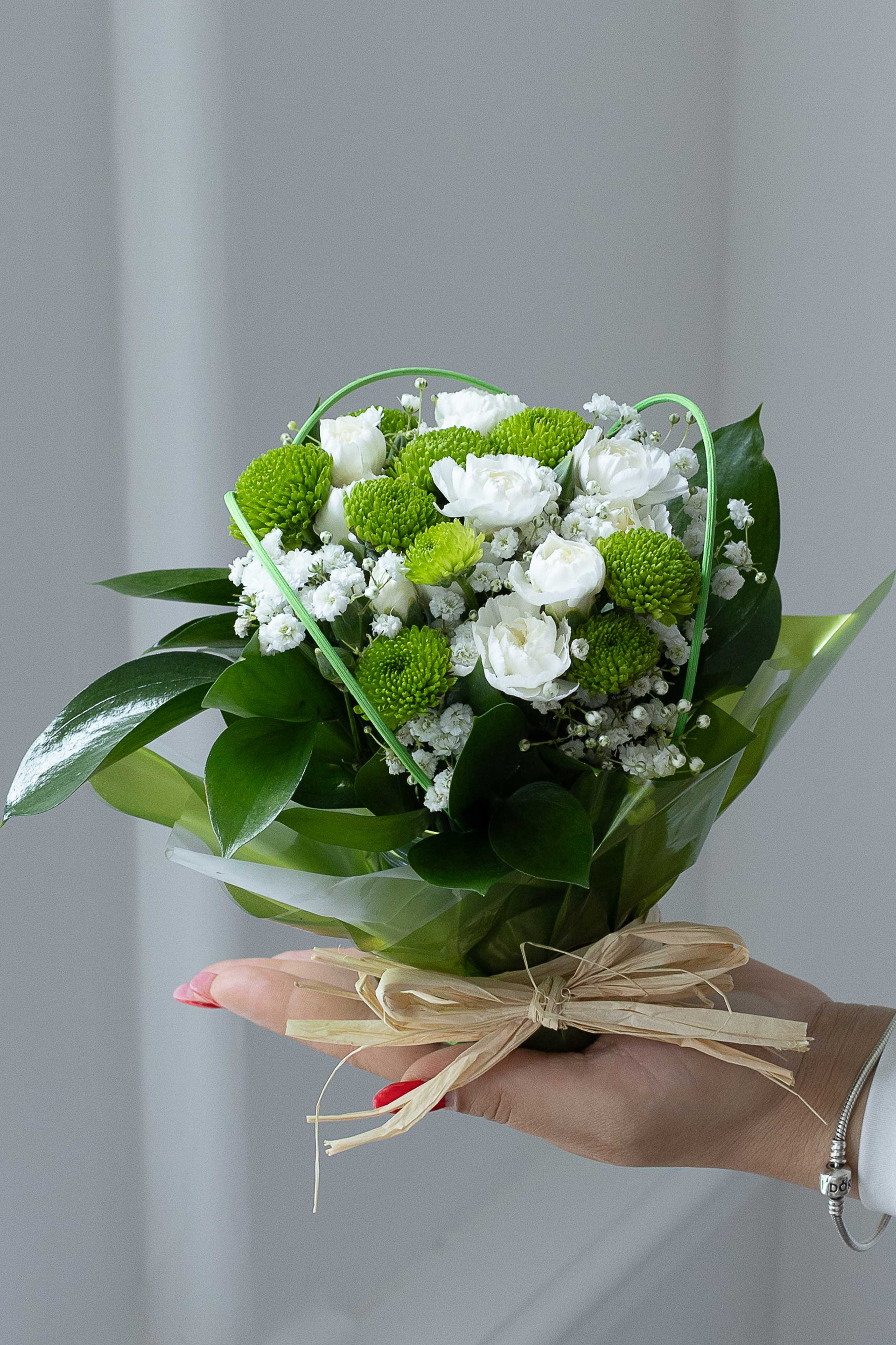 Hand holding a posy of green button chrysanthemums, white lisianthus, and baby's breath wrapped in green paper.
