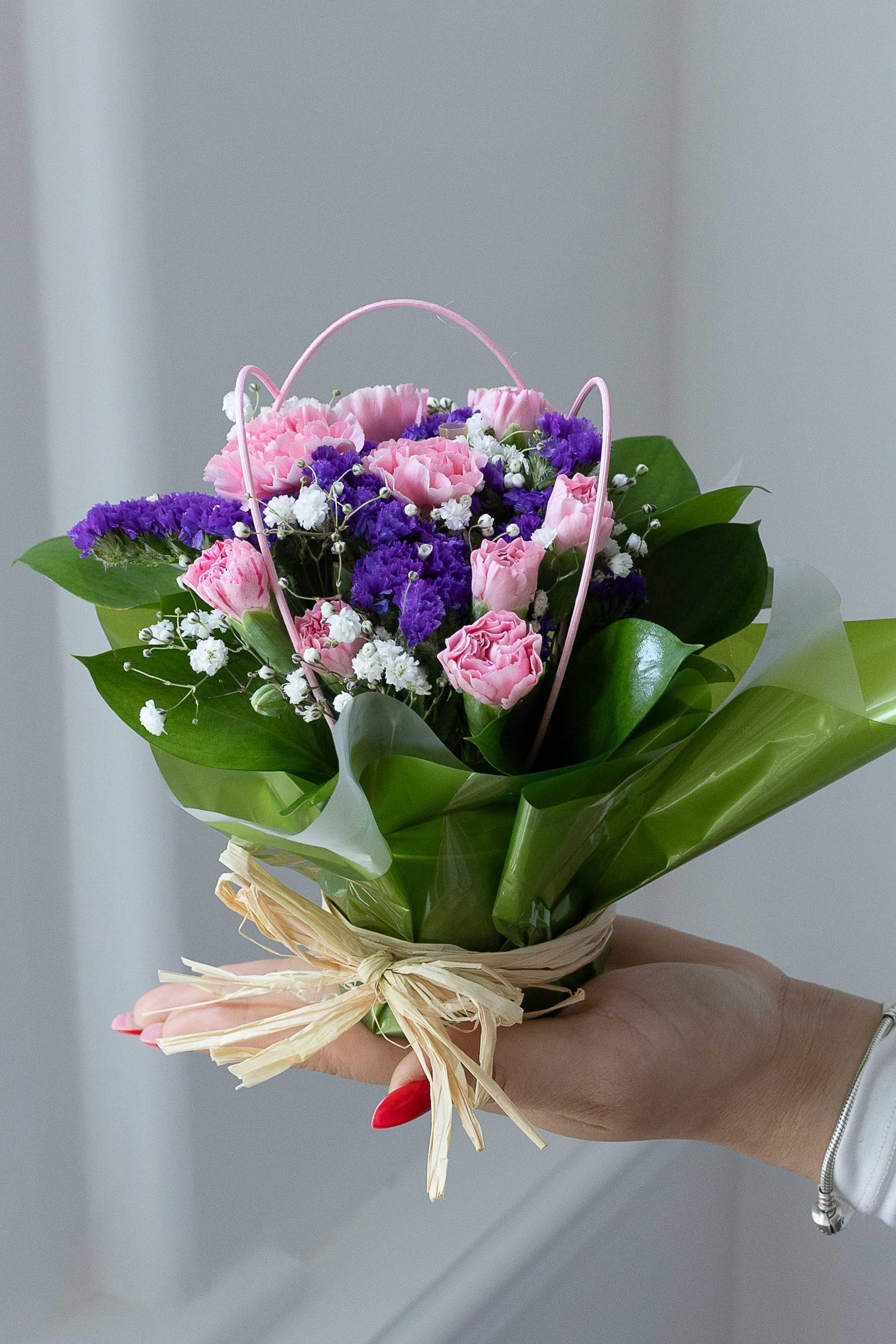 Hand holding a posy of pink carnations, purple statice, and white baby's breath wrapped in green paper and tied with raffia.