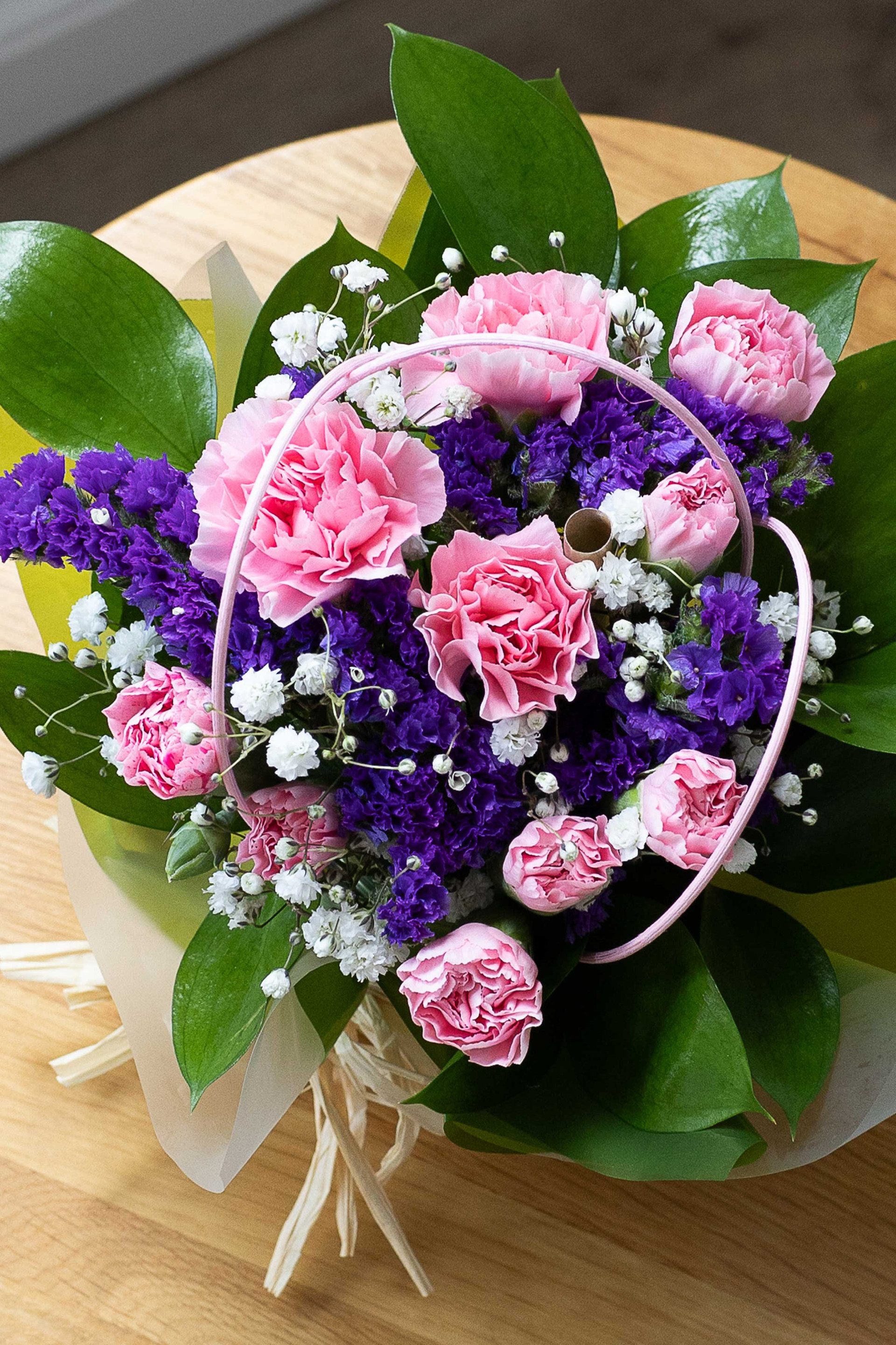 Top-down view of a posy of pink carnations, purple statice, and white baby's breath with green foliage on a wooden table.