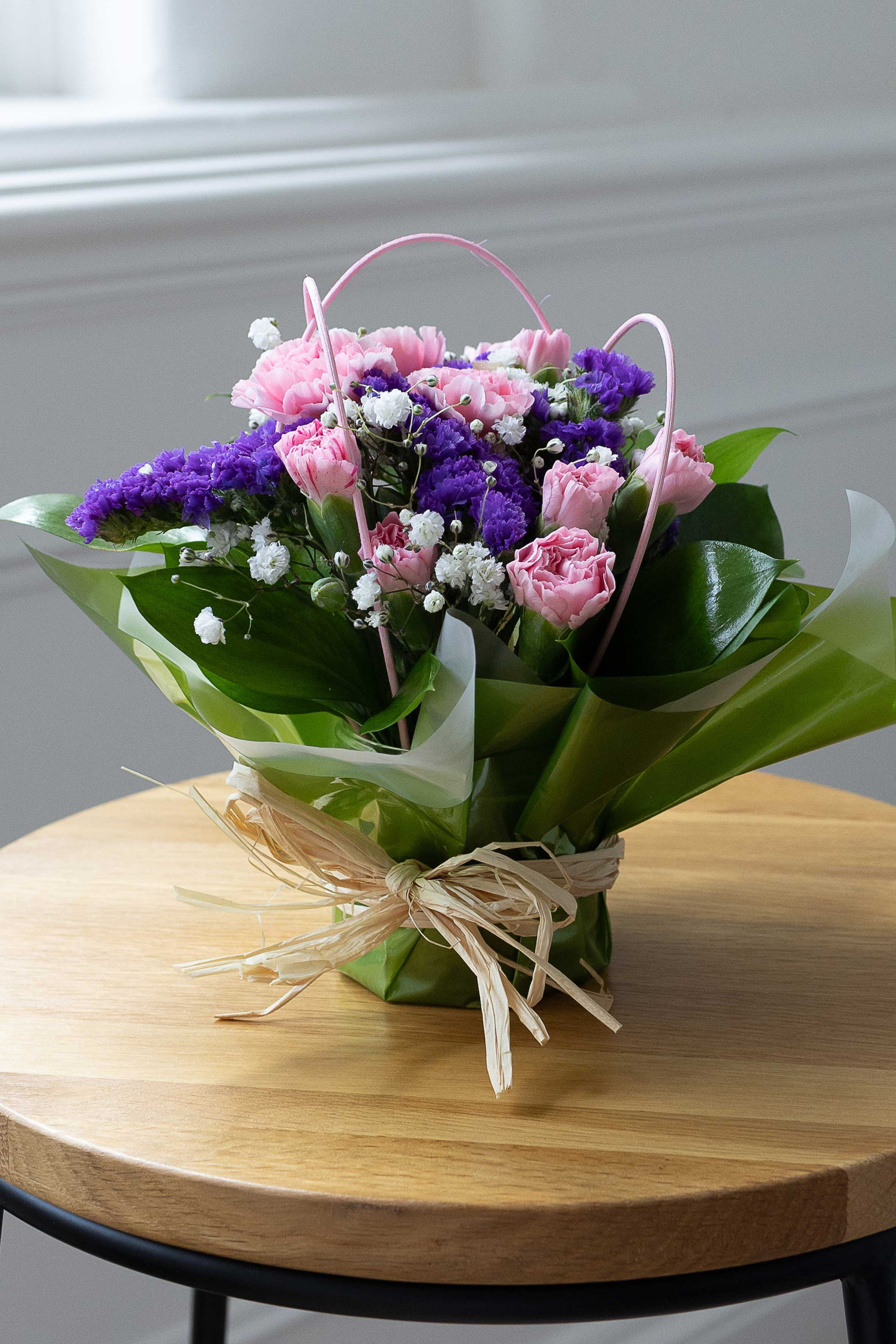 Pink carnations, purple statice, and white baby's breath posy wrapped in green and white paper, displayed on a wooden table.