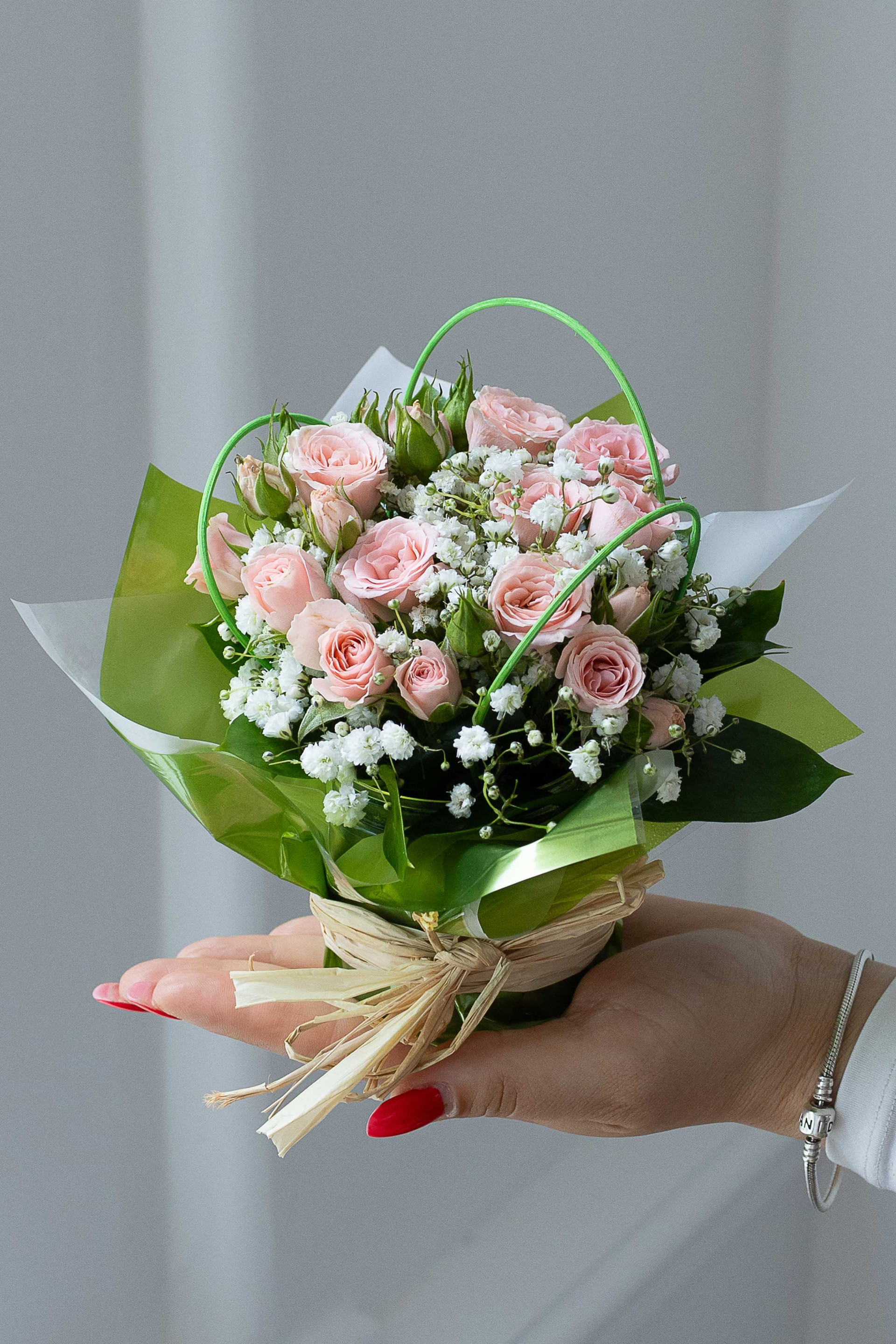 Hand holding a small posy of pale pink roses and white baby's breath wrapped in green paper and tied with raffia.