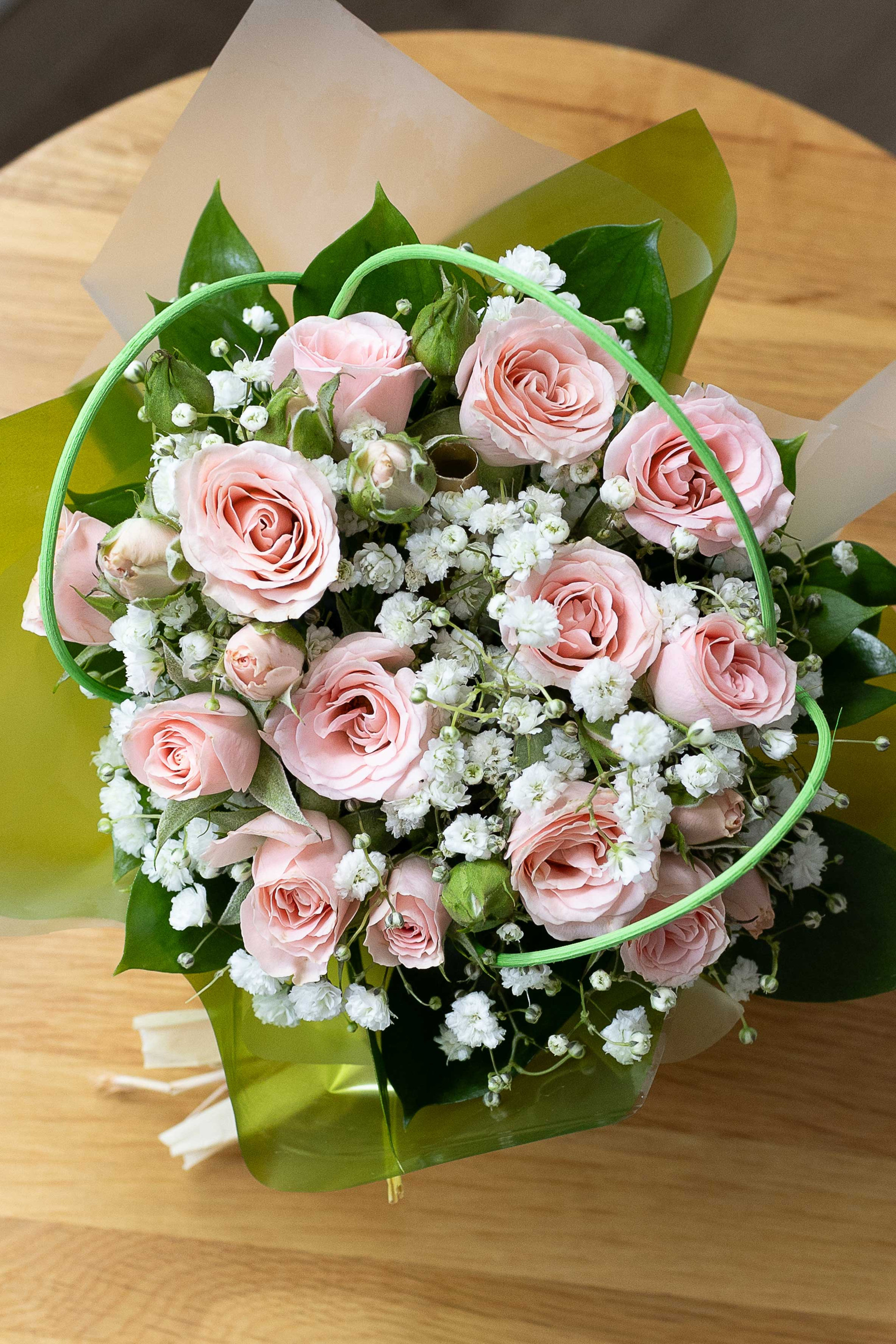 Top-down view of a posy of pale pink roses and white baby's breath wrapped in green and white paper on a wooden table.