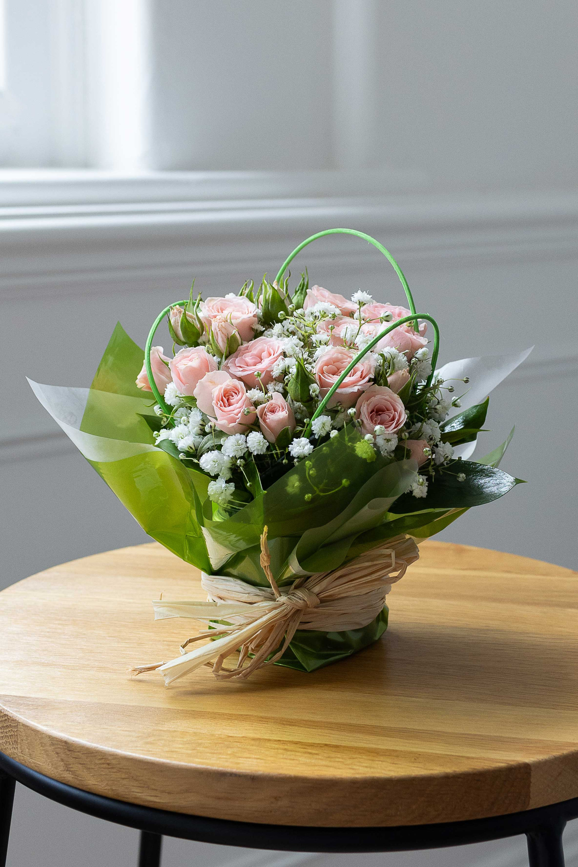 Pink rose and baby's breath posy wrapped in green and white paper, displayed on a round wooden table.