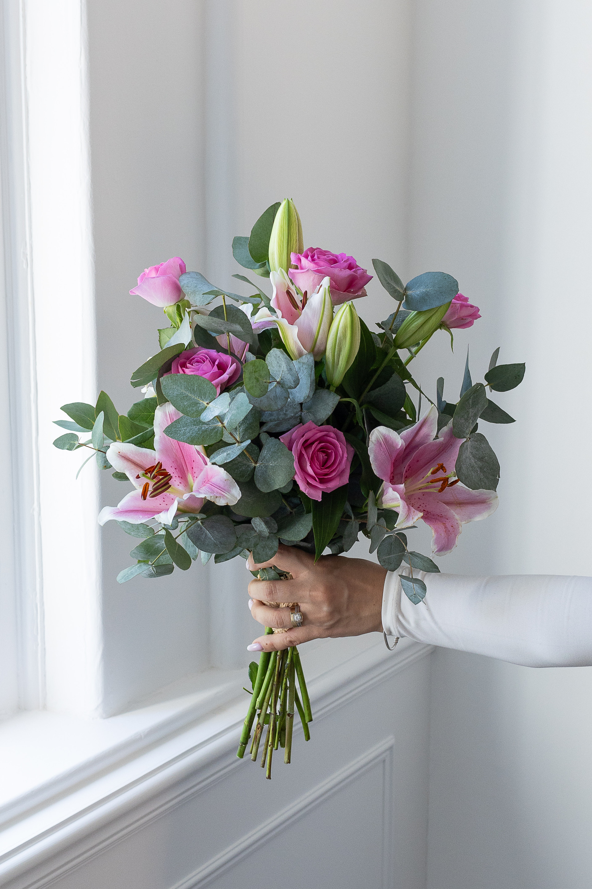 Hand holding a bouquet of pink roses, pink lilies, and eucalyptus beside a bright window.