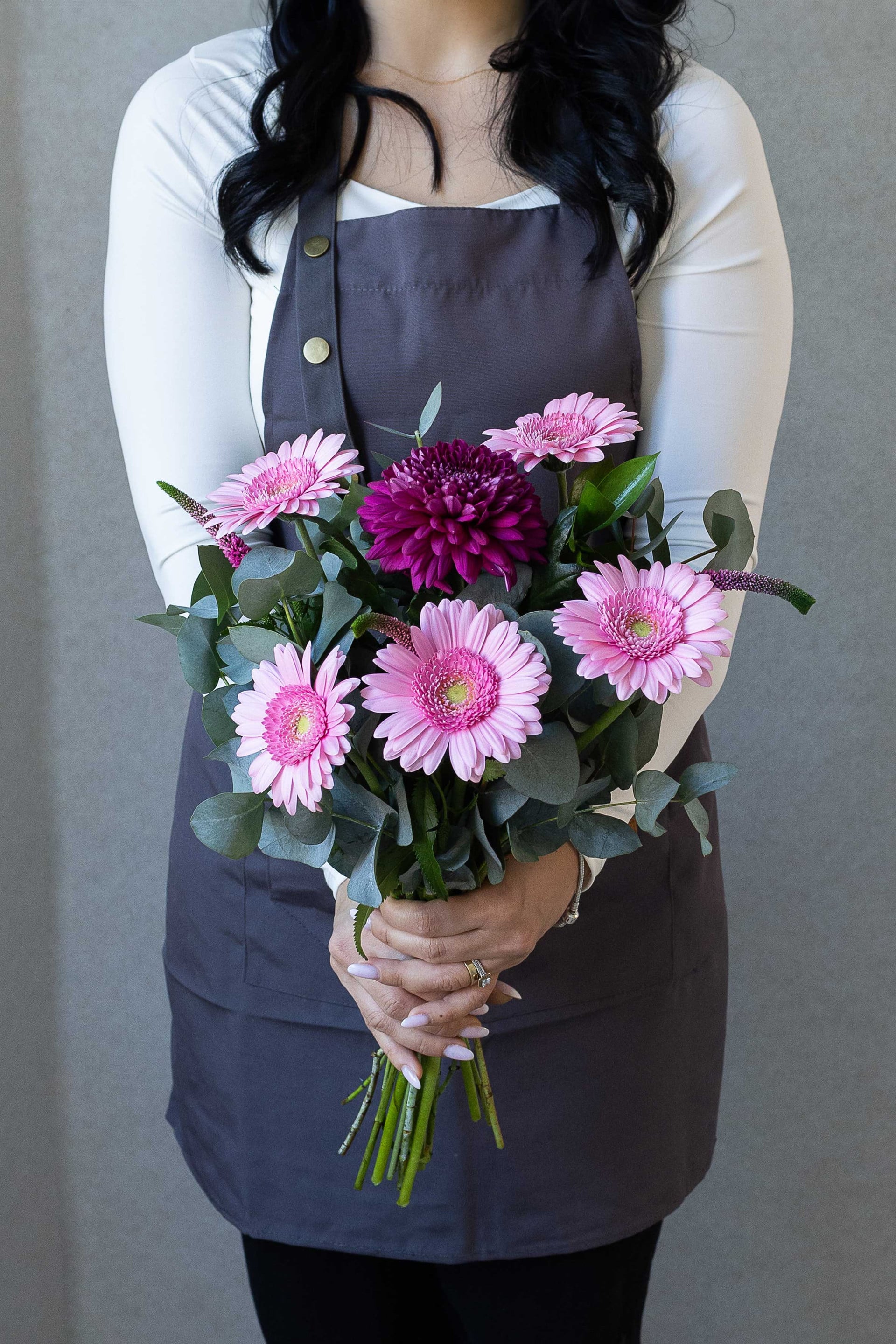 Florist holding a bouquet of pink gerbera daisies, a burgundy chrysanthemum, eucalyptus, and veronica flowers against a gray background.