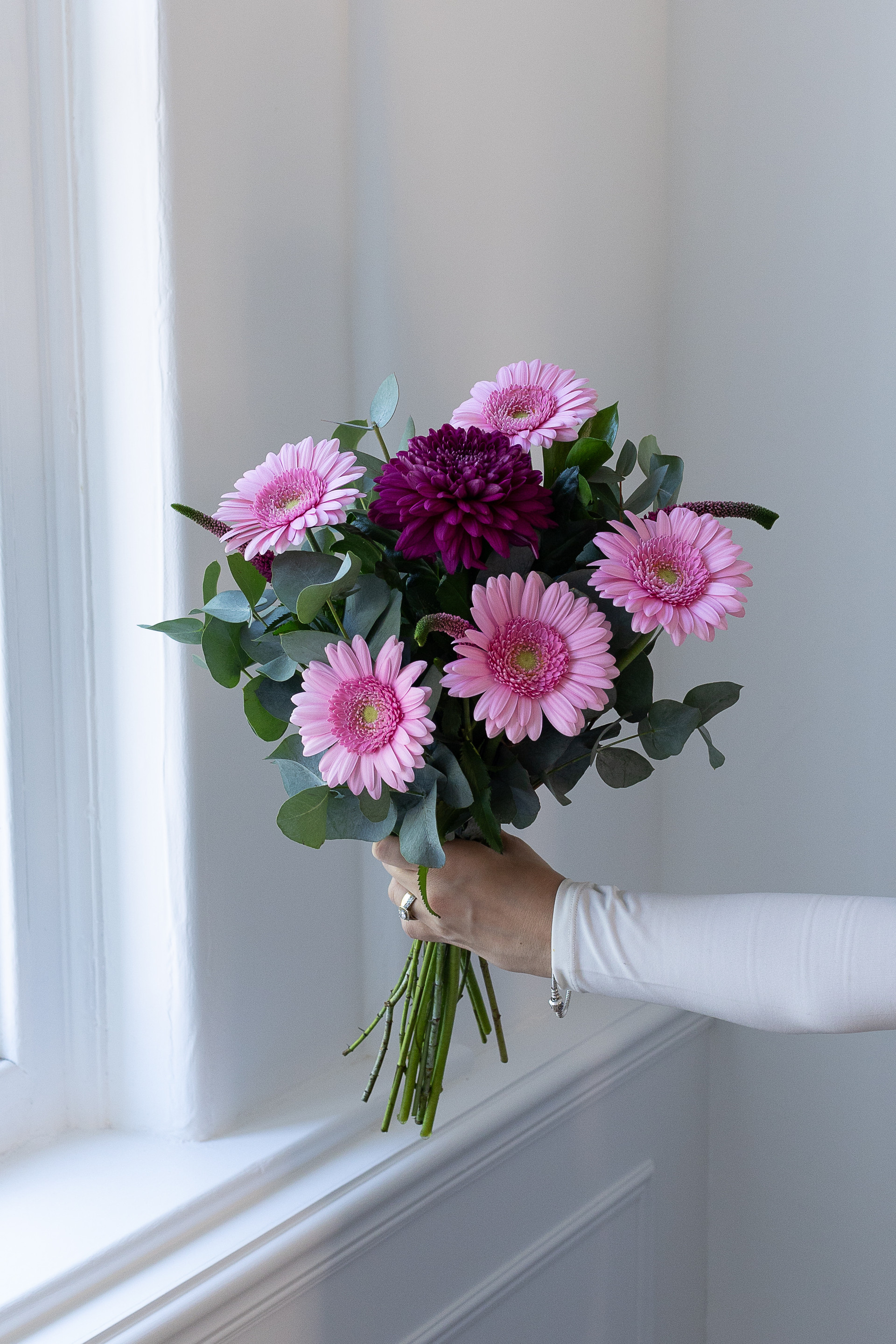 Hand holding a bouquet of pink gerbera daisies, a burgundy chrysanthemum, eucalyptus, and veronica flowers by a window.
