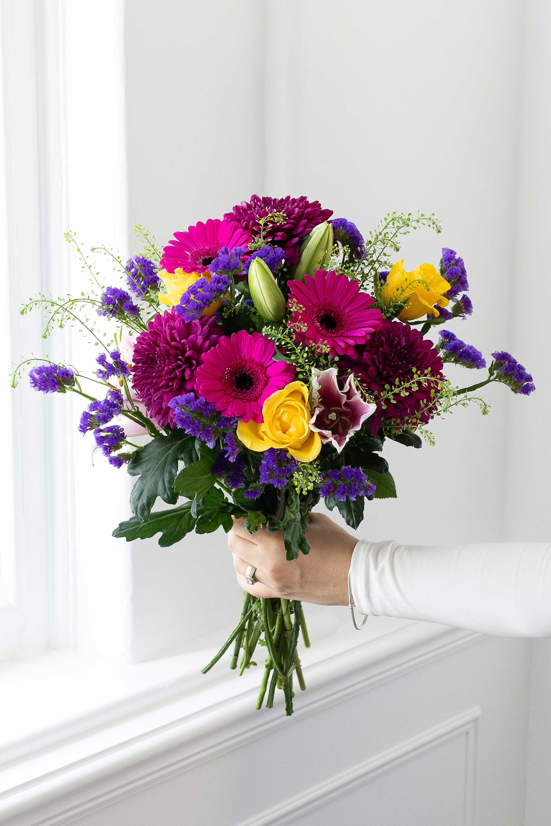 Hand holding a colourful bouquet of gerberas, chrysanthemums, roses, lilies, and statice by a window.