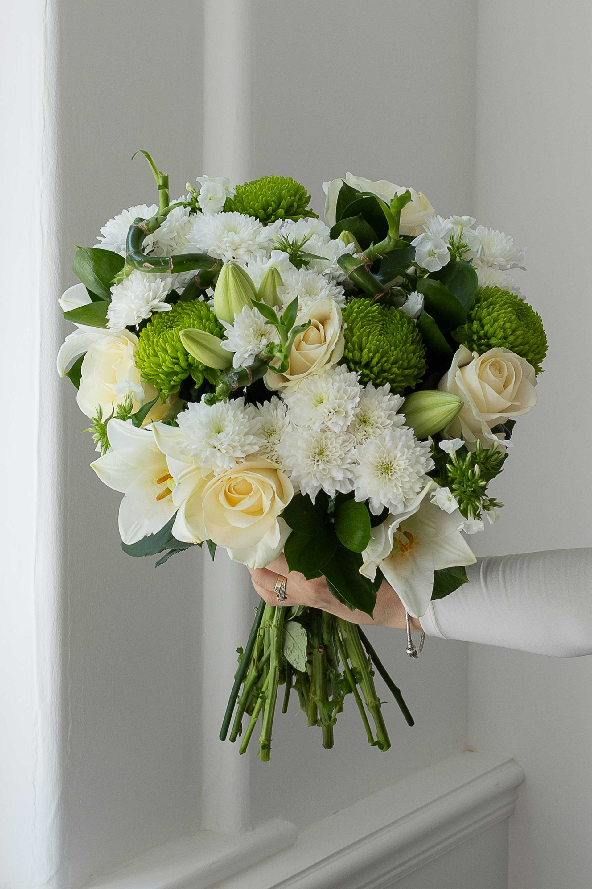 White and green bouquet held by a window with roses, lilies, and chrysanthemums.