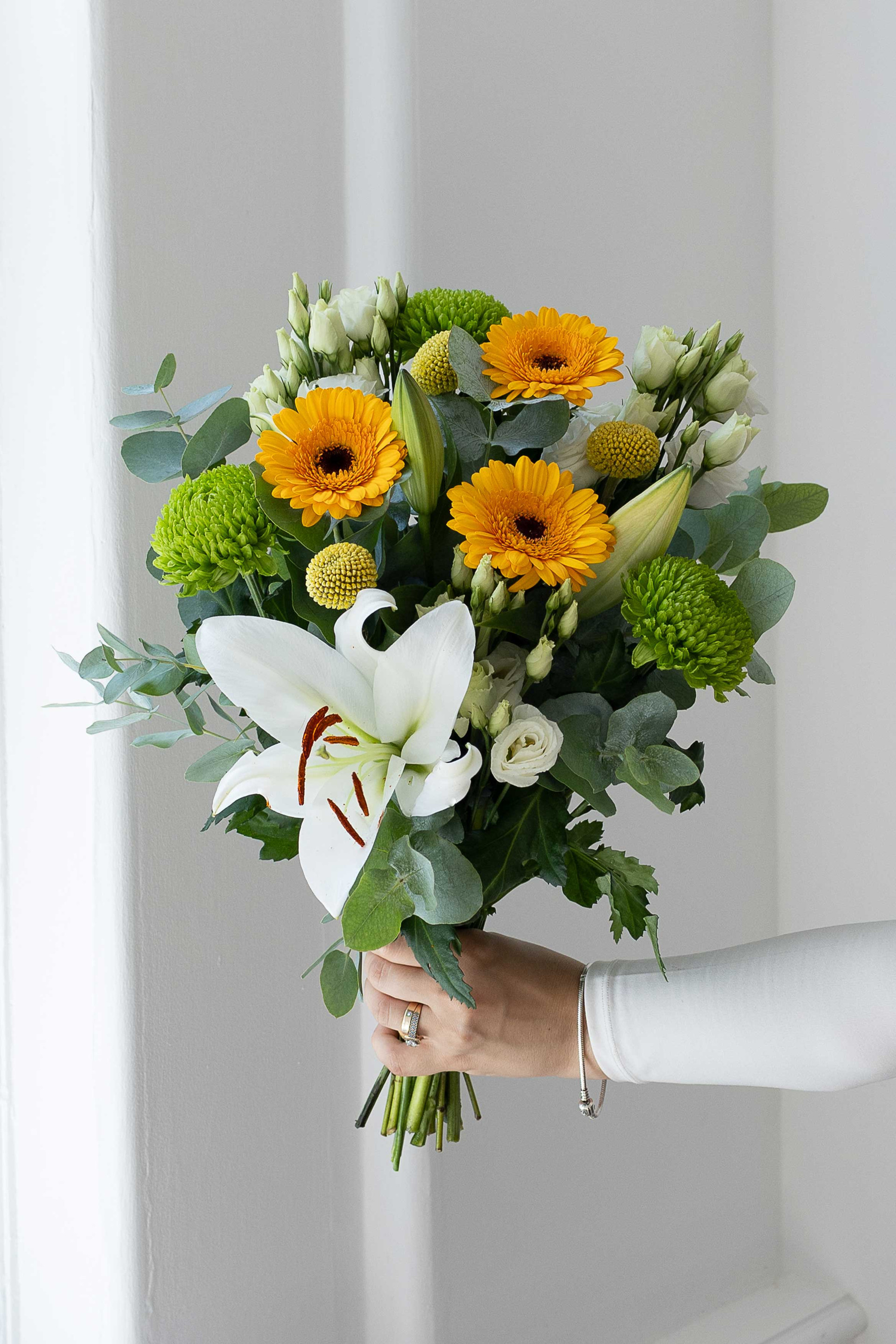 Hand holding a yellow, white, and green bouquet by a window.
