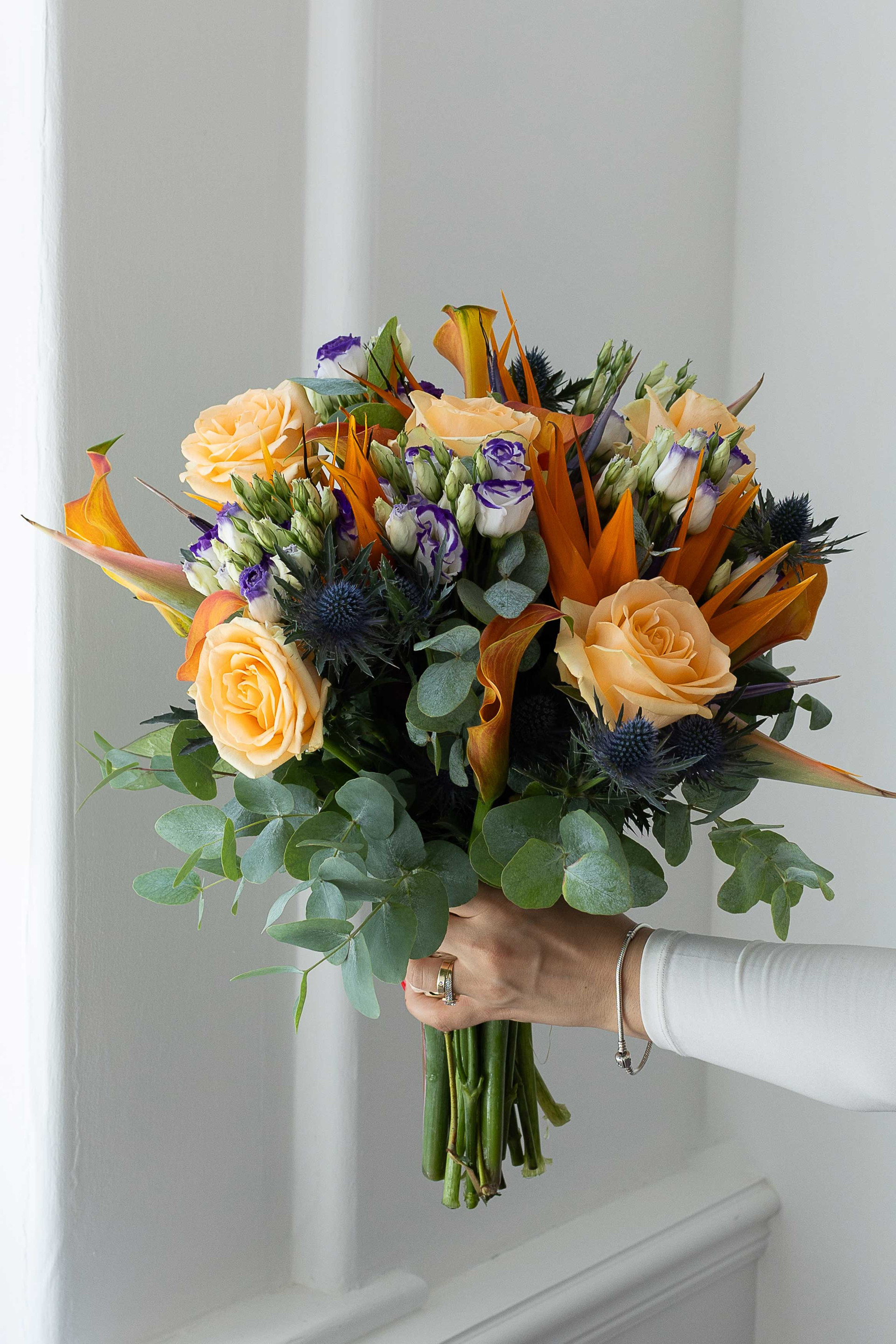 Hand holding a peach rose and bird of paradise bouquet by a window.