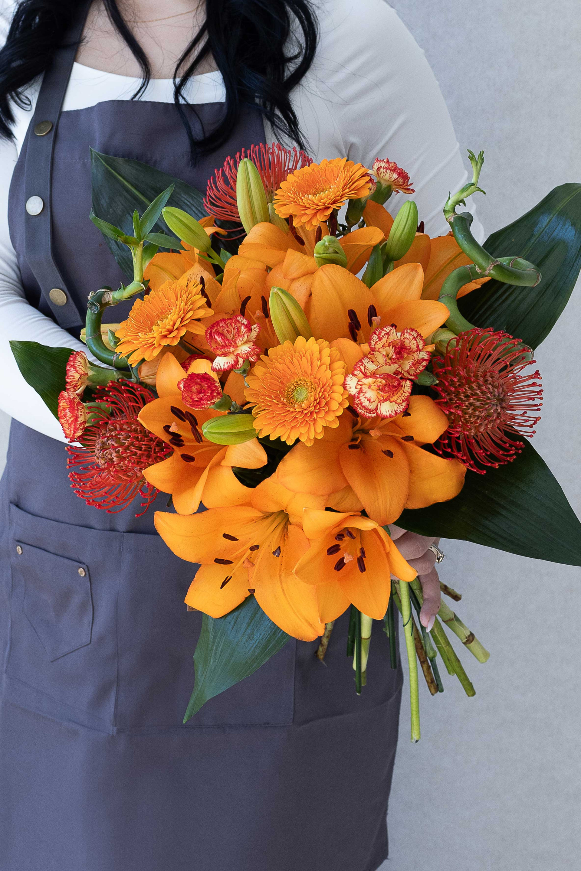 Orange lily bouquet with gerberas, proteas, carnations, and decorative bamboo stems.