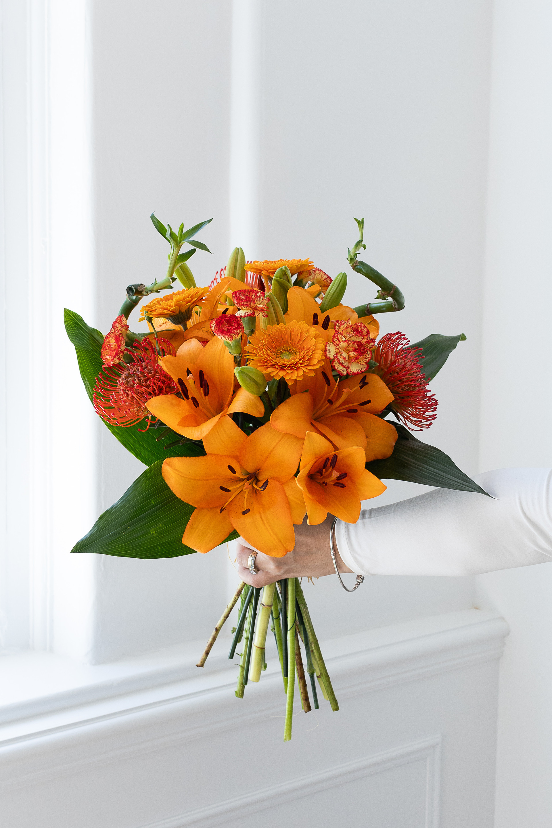 Hand holding a bright bouquet of orange lilies, gerberas, proteas, and carnations by a window.