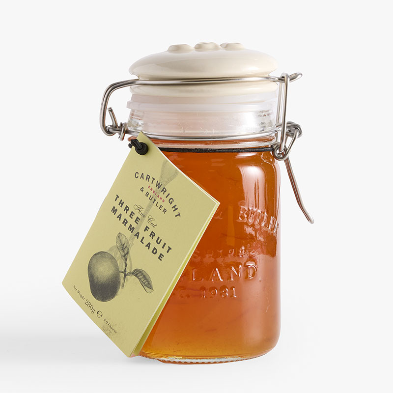 Clip-top glass jar of Cartwright & Butler Three Fruit Marmalade with a pale yellow hanging label and amber-orange marmalade visible inside.