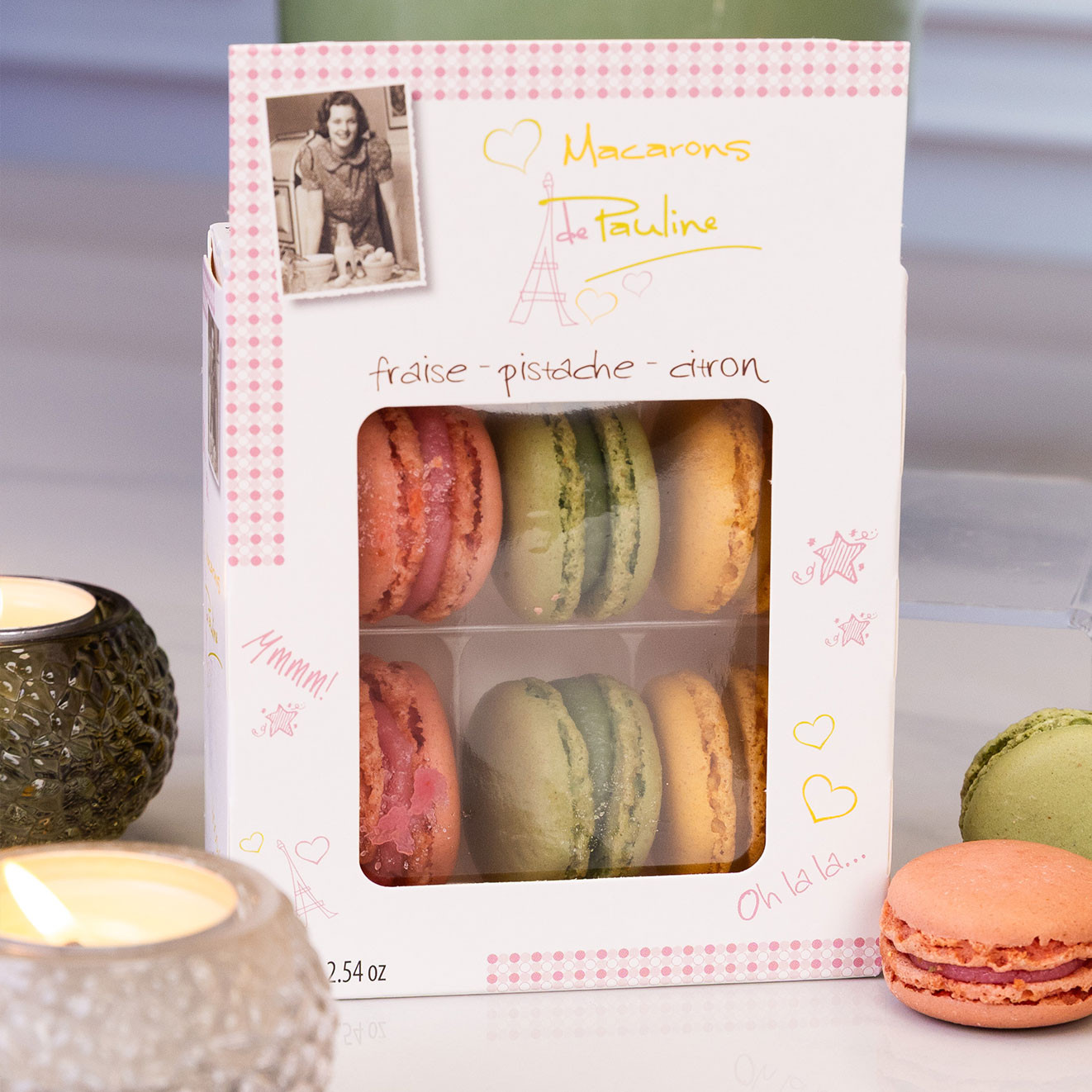 Box of Macarons de Pauline French macarons in strawberry, pistachio and lemon flavours displayed with loose macarons and lit candles.