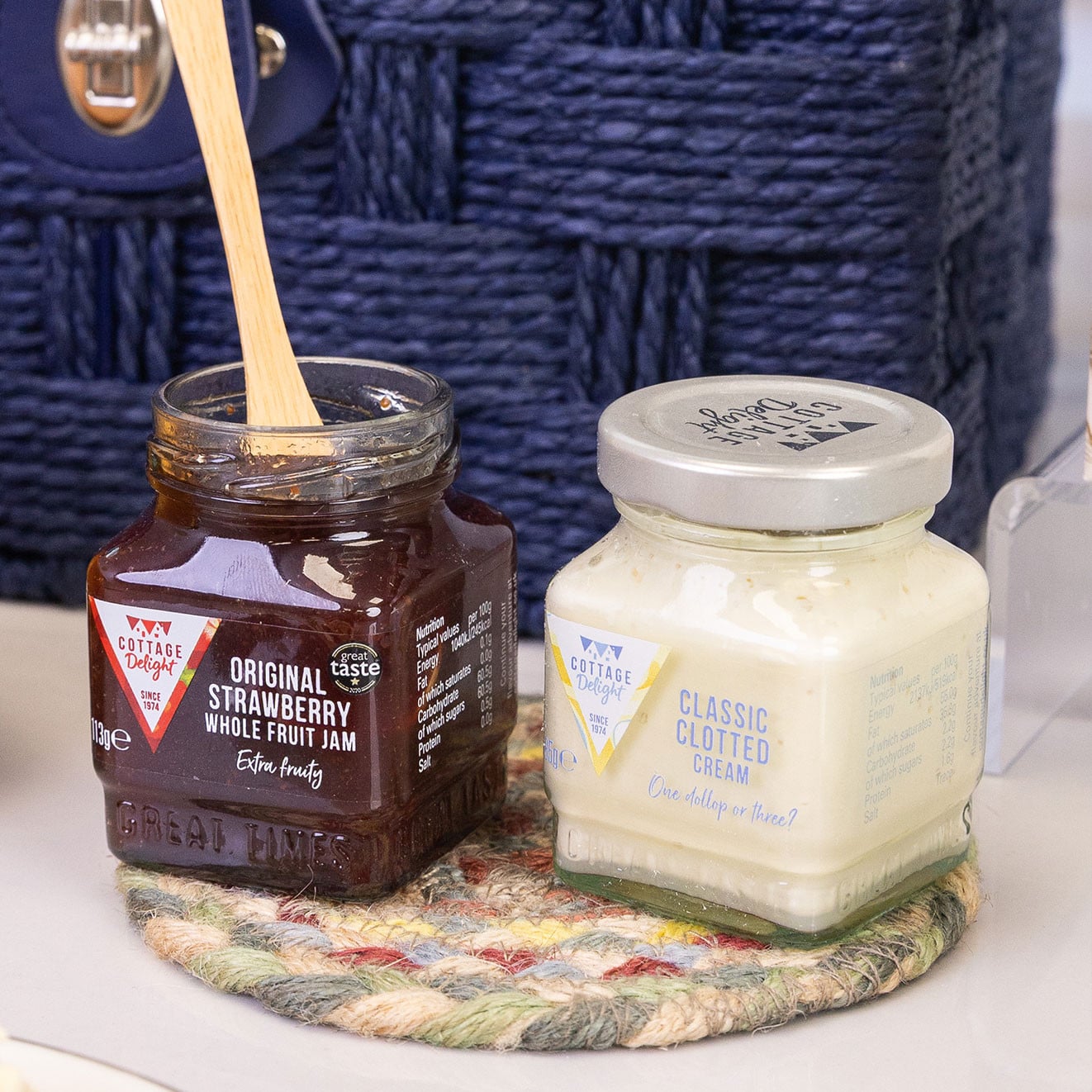 Jars of Cottage Delight Original Strawberry Whole Fruit Jam and Classic Clotted Cream displayed beside a hamper.