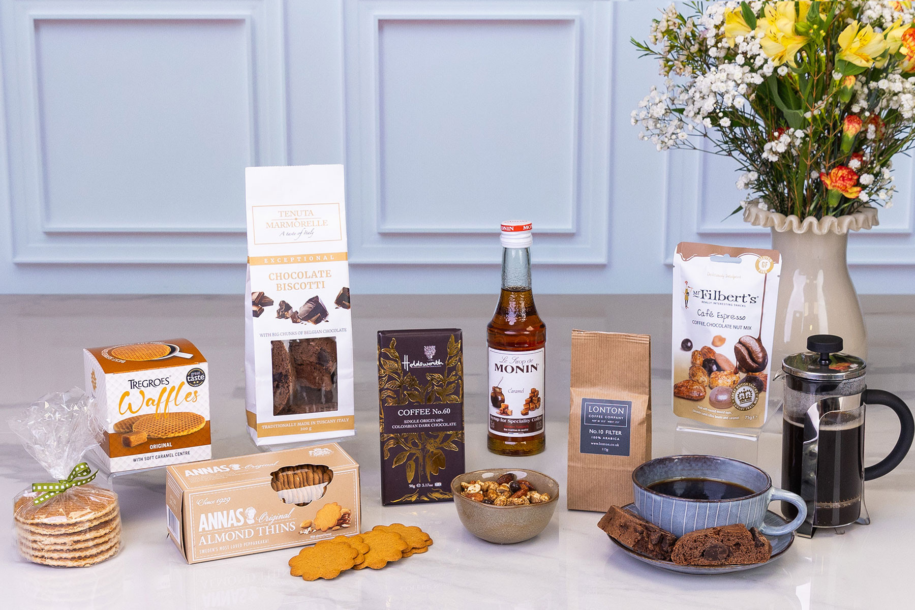 Luxury coffee gift hamper featuring ground coffee, French press, chocolate biscotti, coffee syrup, caramel waffles, almond biscuits, dark chocolate, coffee and chocolate nut mix, and fresh flowers.