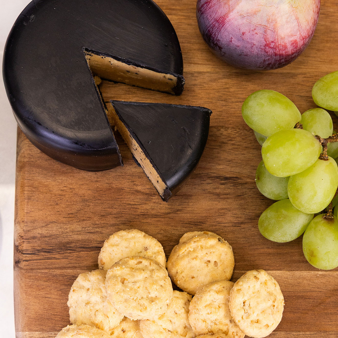 Round smoked cheese with sliced wedge served on a wooden board alongside savoury biscuits and fresh green grapes.