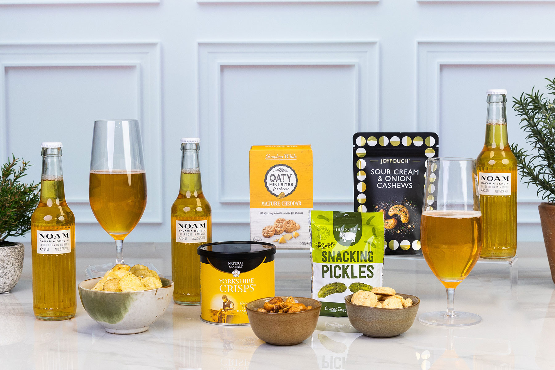 Beer and savoury snacks gift hamper featuring NOAM lager, Yorkshire Crisps, Grandma Wild's oat biscuits, Serious Pig Snacking Pickles, sour cream and onion cashews, and mixed nuts.