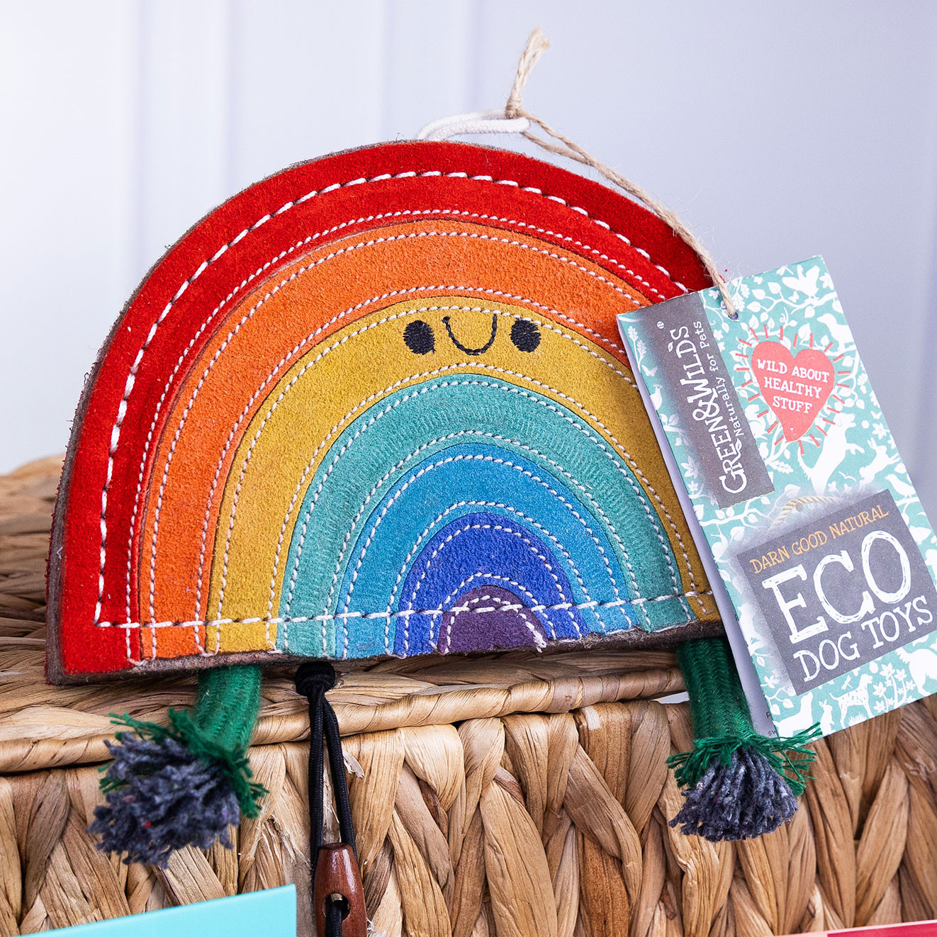 Colourful rainbow-shaped Green & Wild's eco dog toy displayed on top of a woven basket with product tag attached.