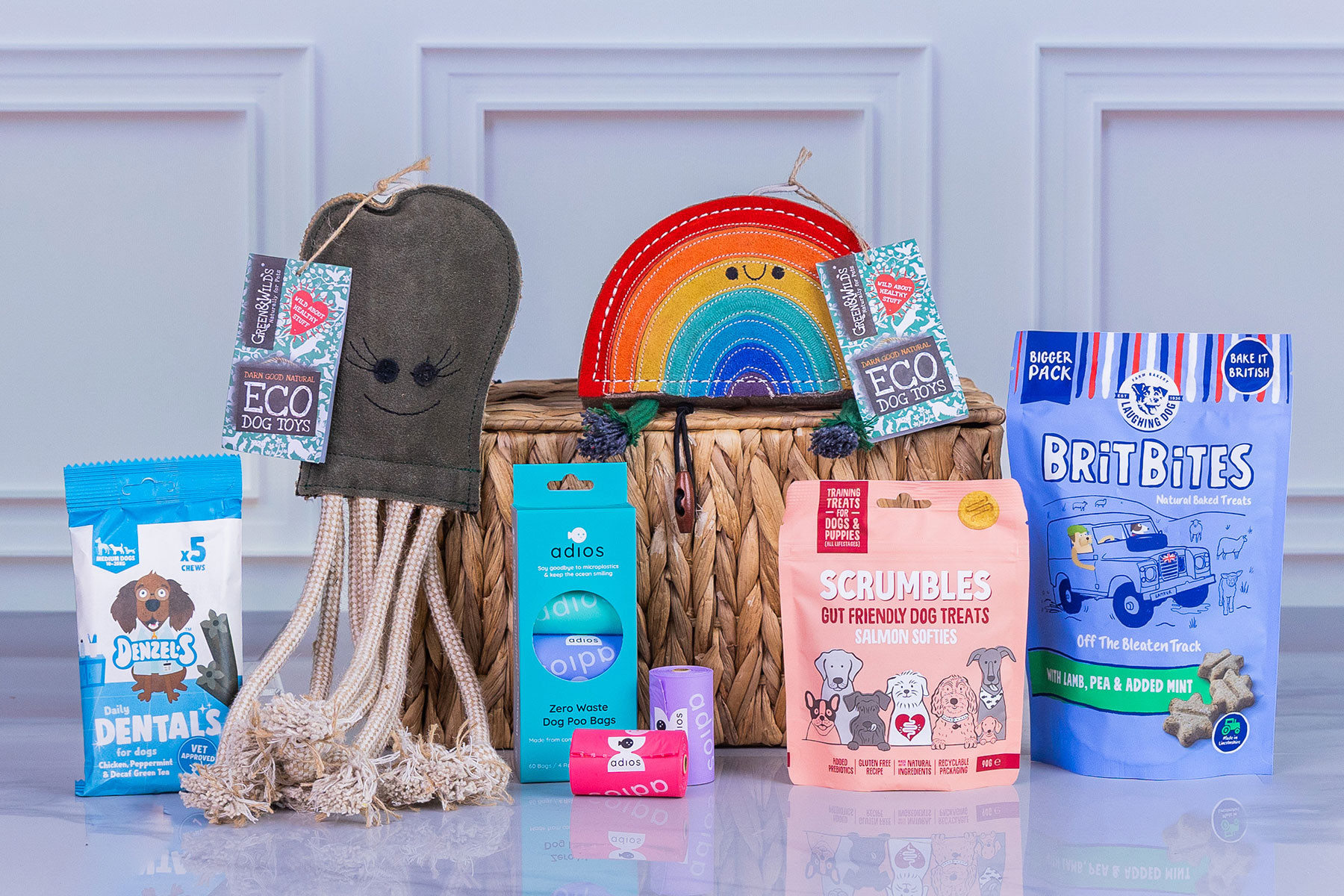 Selection of eco-friendly dog products including natural treats, dental chews, compostable dog poo bags, and sustainable dog toys displayed in front of a woven storage basket.