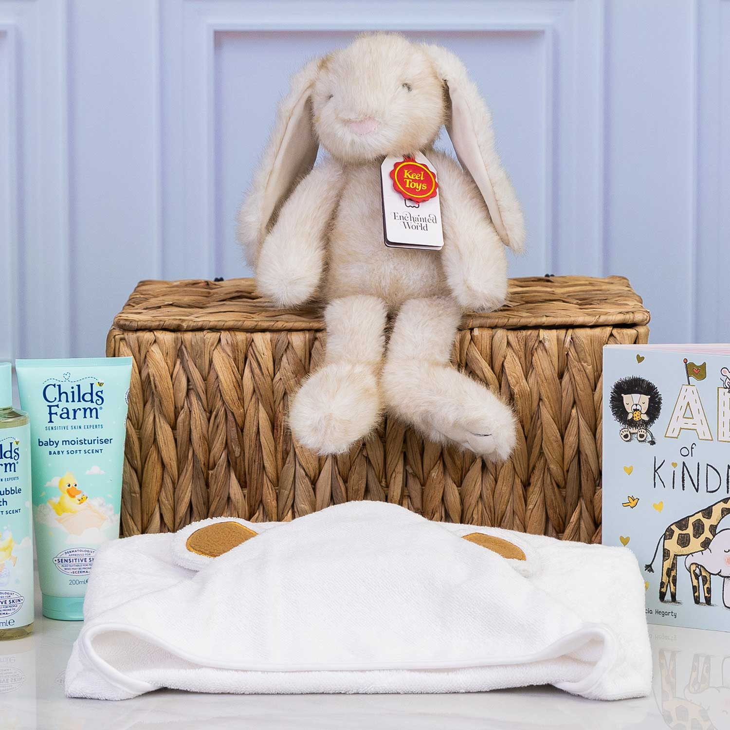 Close up of Luxury baby gift hamper featuring Childs Farm baby toiletries, a soft plush bunny, ABC of Kindness board book and a hooded baby towel displayed around a wicker hamper.