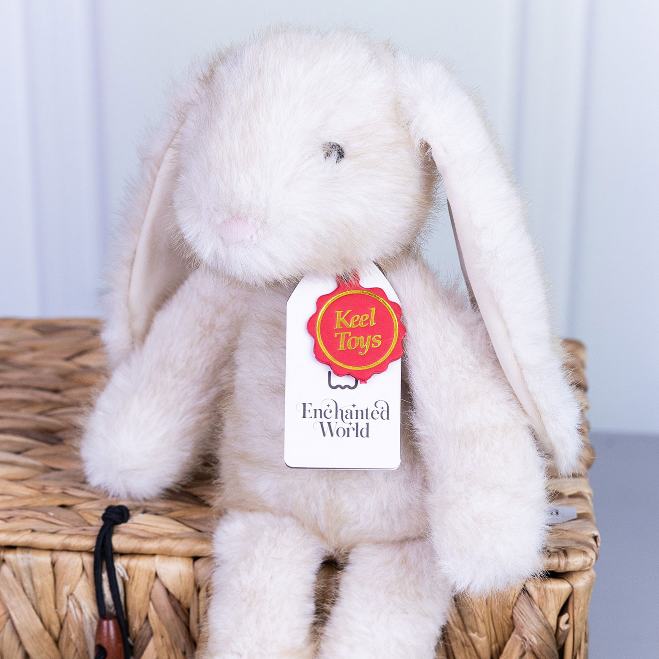 Soft cream plush bunny from the Keel Toys Enchanted World collection sitting on a wicker hamper with branded swing tag attached.
