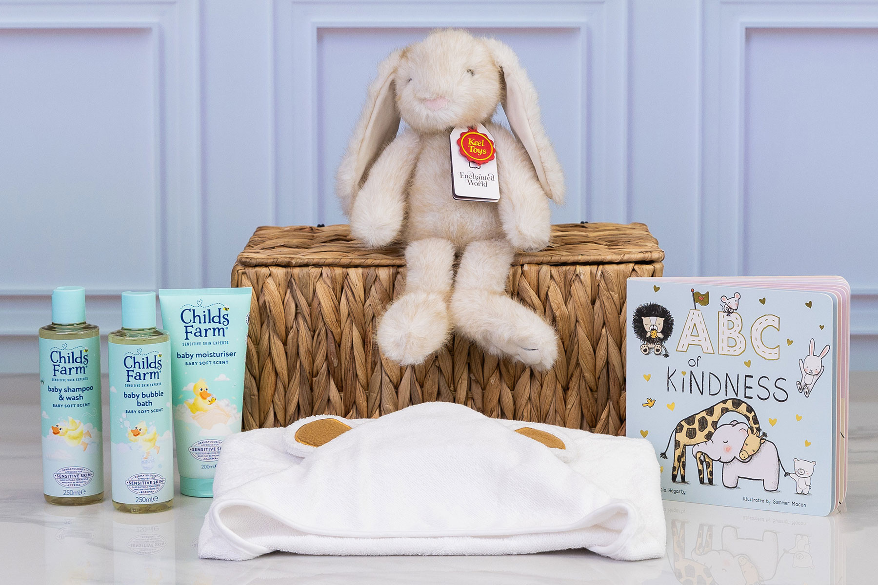 Luxury baby gift hamper featuring Childs Farm baby toiletries, a soft plush bunny, ABC of Kindness board book and a hooded baby towel displayed around a wicker hamper.