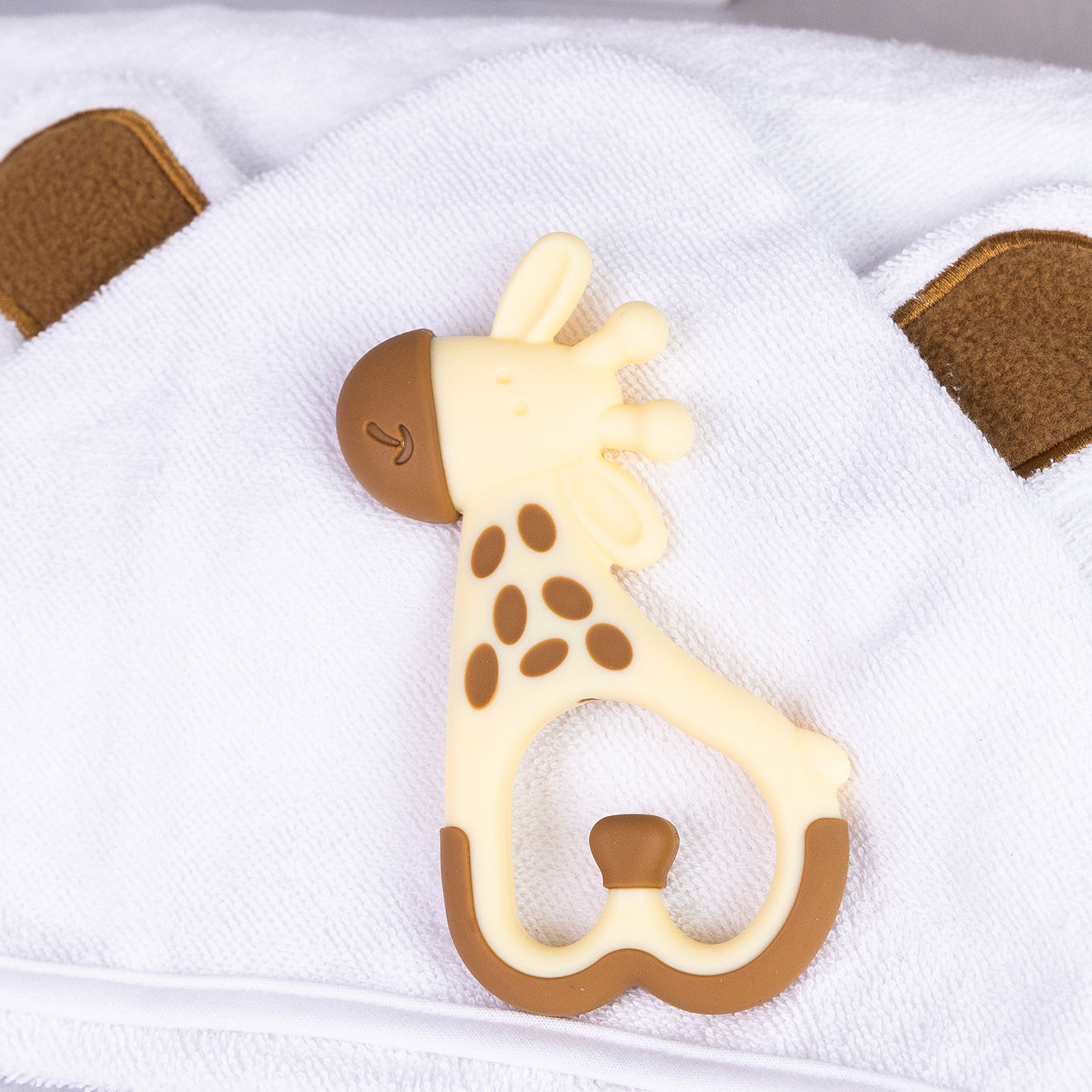 Cream and beige giraffe-shaped silicone baby teether resting on a soft white hooded baby towel with bear ears.