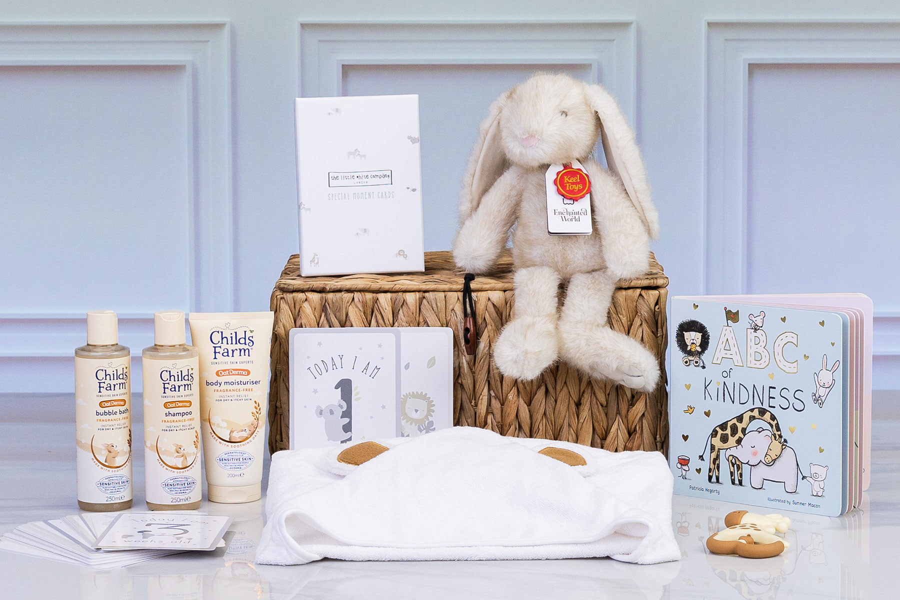 Luxury baby gift hamper featuring Childs Farm OatDerma skincare, a plush bunny, ABC of Kindness board book, milestone cards, hooded baby towel and giraffe-shaped silicone teether.