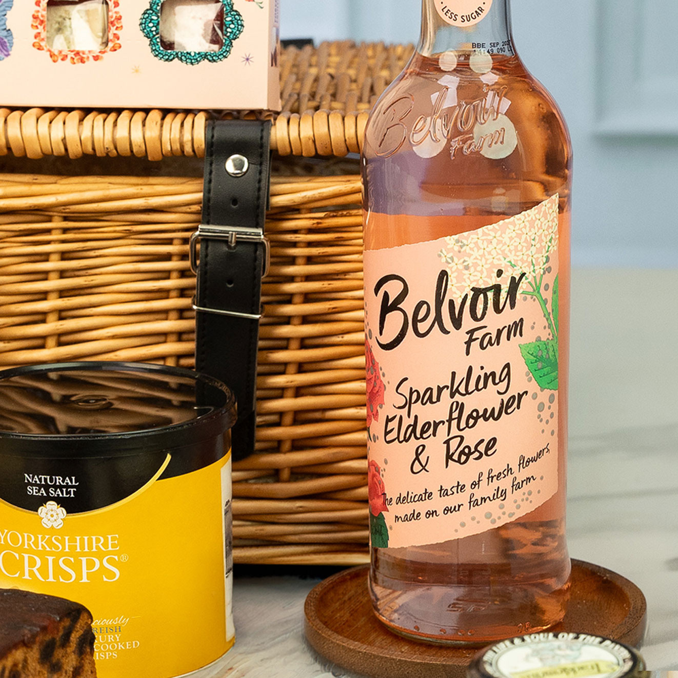 Bottle of Belvoir Farm Sparkling Elderflower & Rose drink displayed alongside Yorkshire Crisps and a traditional willow hamper.