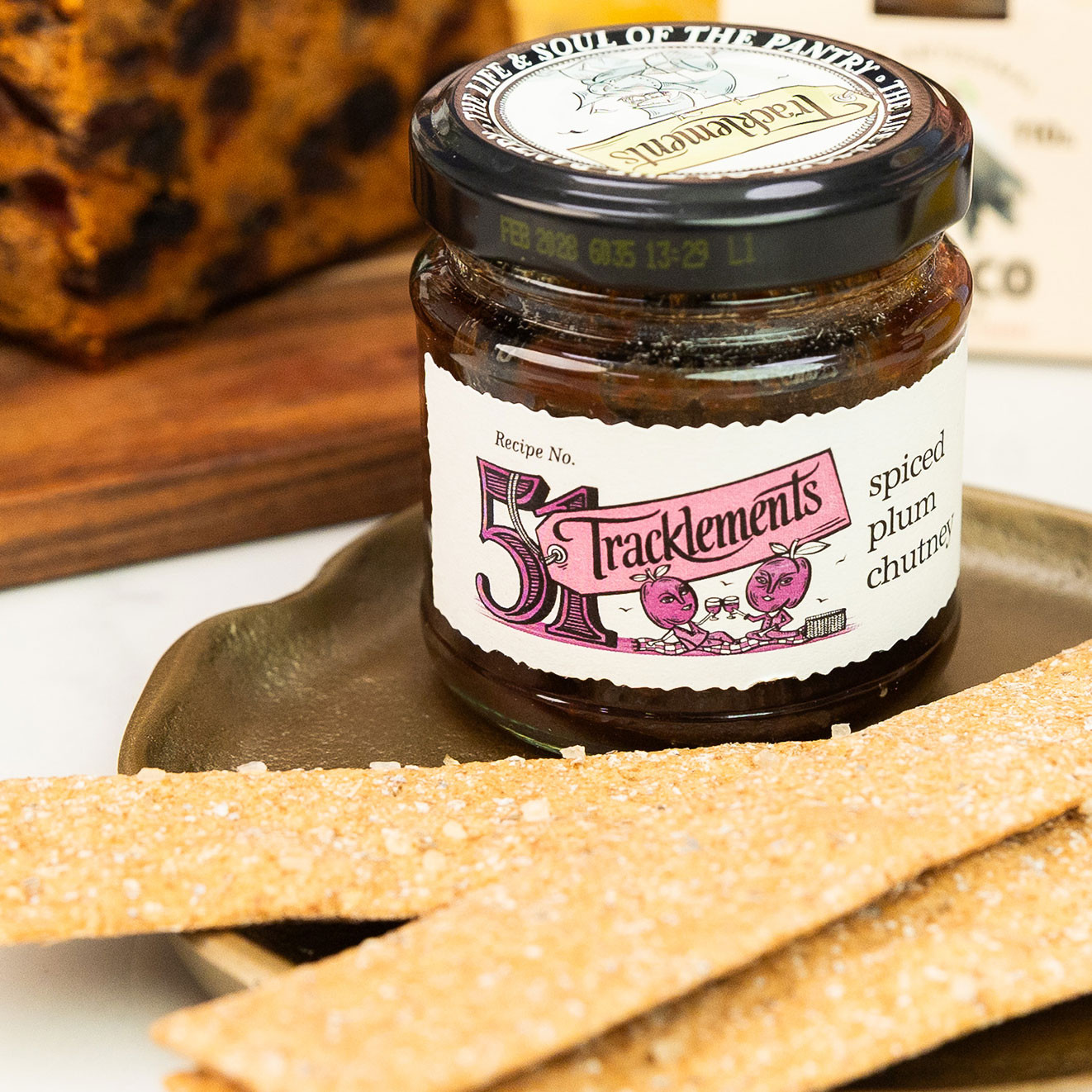 Jar of Tracklements Spiced Plum Chutney served with crispbread crackers, with fruit cake and gourmet foods in the background.
