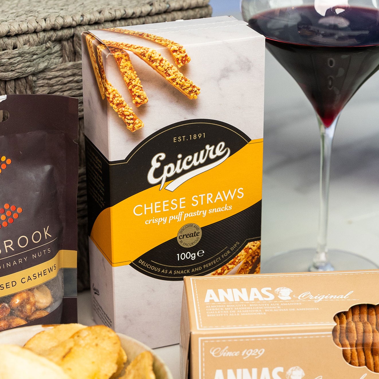 Box of Epicure Cheese Straws displayed with a glass of red wine, Anna's Almond Thins and gourmet snacks.