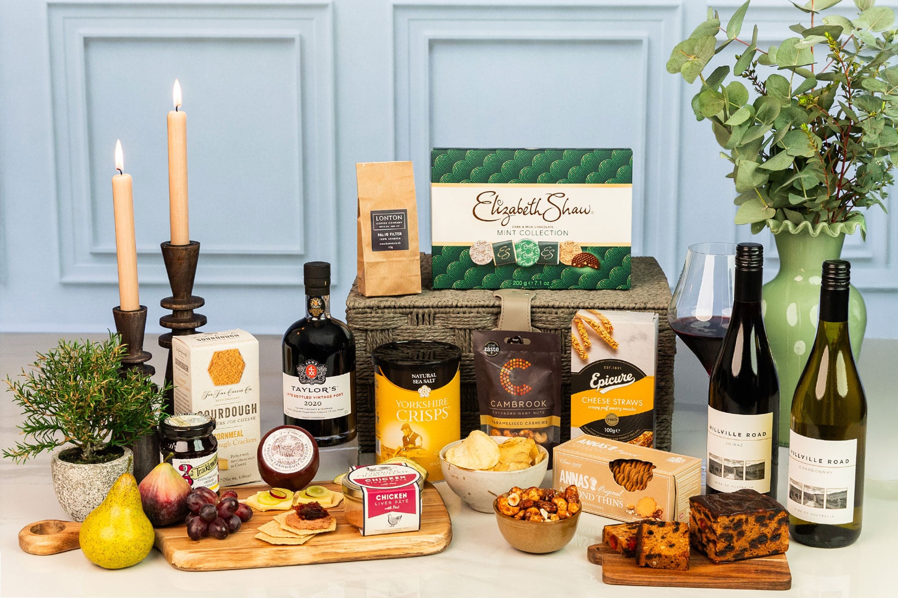 Luxury wine and gourmet hamper with red and white wine, artisan cheeses, savoury snacks, chocolates, fruit cake, port and premium pantry treats arranged around a grey rope hamper.