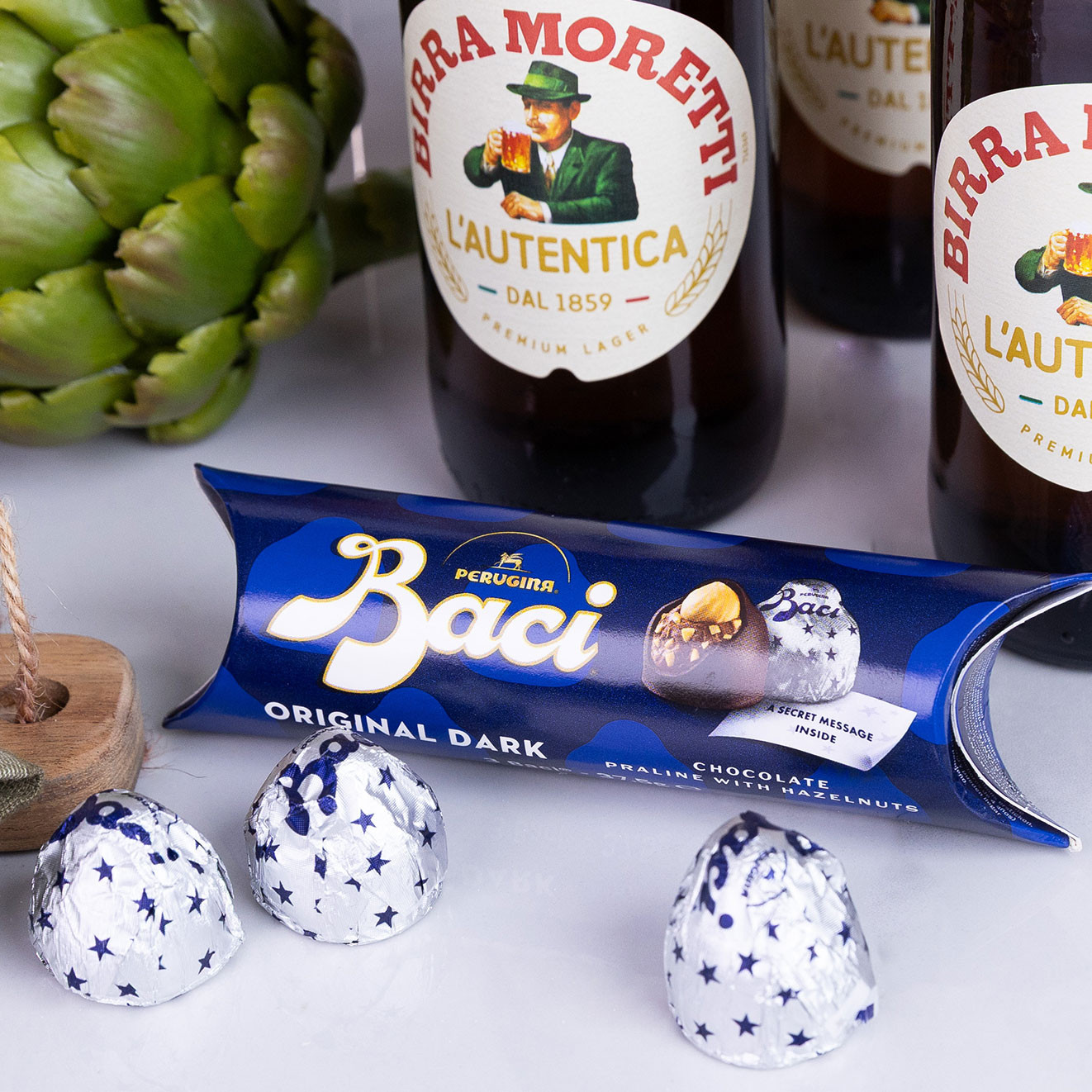Perugina Baci Original Dark chocolate pralines with hazelnuts displayed alongside Birra Moretti lager bottles and fresh artichokes.