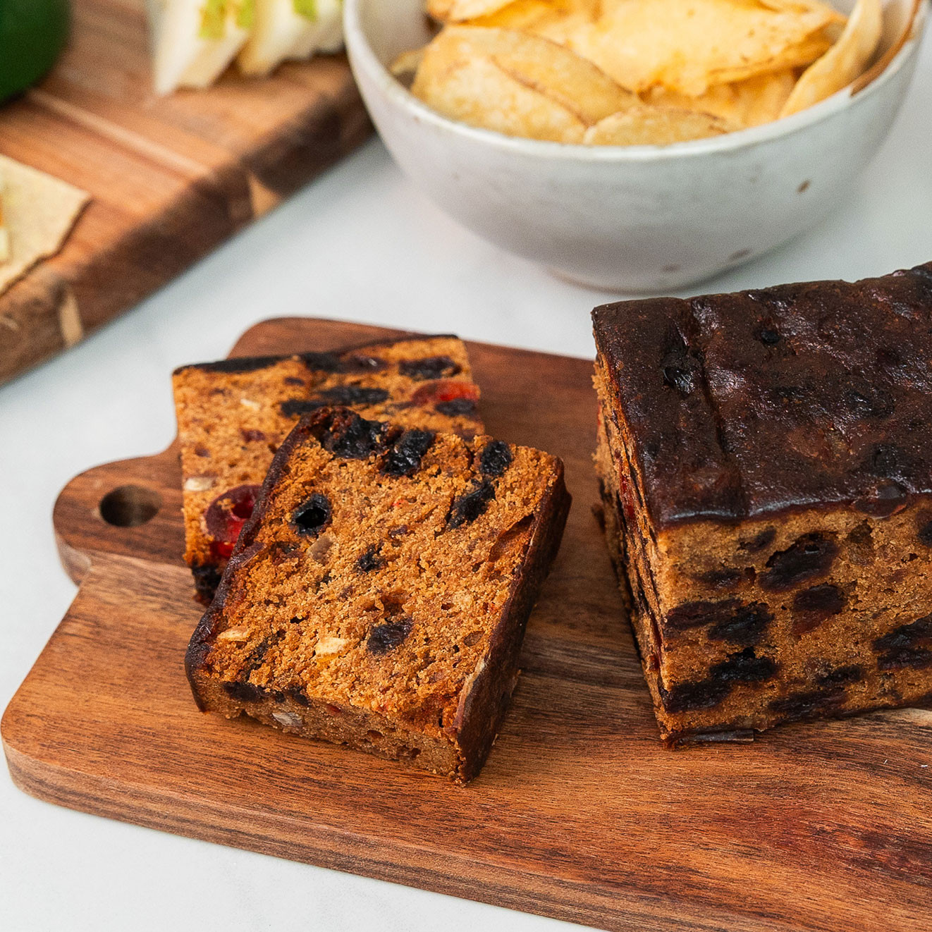 Rich sliced fruit cake displayed on a wooden serving board with gourmet crisps and artisan cheese in the background.