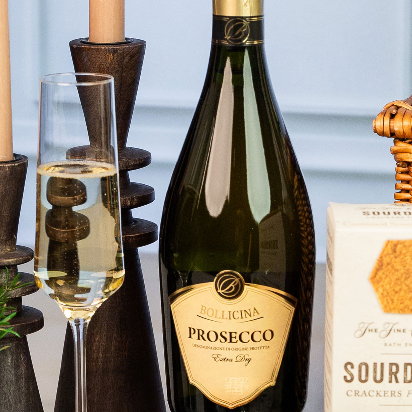 Bottle of Bollicina Prosecco Extra Dry served with a filled champagne flute beside artisan sourdough crackers and lit candles.