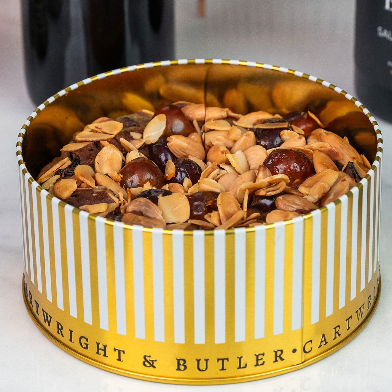 Luxury fruit cake presented in an elegant Cartwright & Butler striped gold gift tin with a generous nut-topped cake inside.