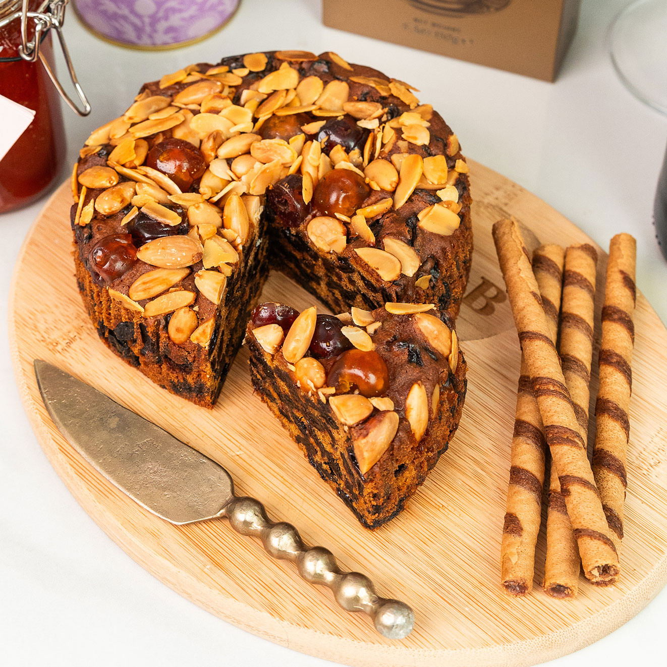 Rich fruit cake topped with whole cherries and toasted almonds, served with chocolate-filled wafer rolls and a decorative cake knife.