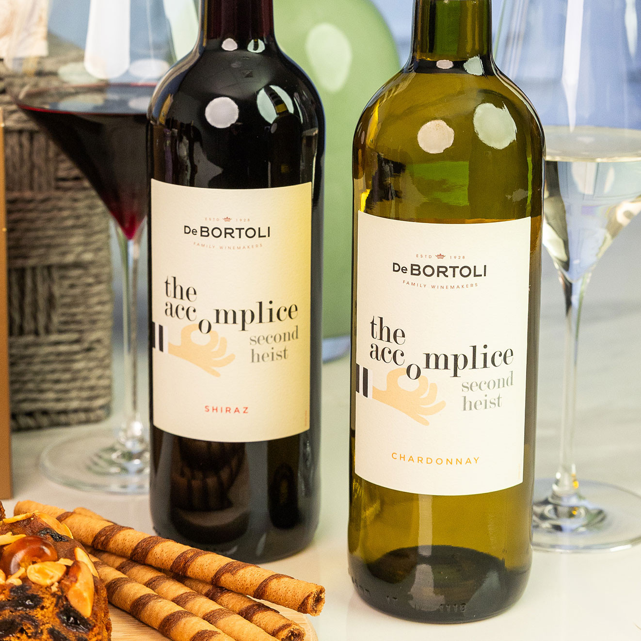 De Bortoli The Accomplice Shiraz and Chardonnay displayed with red and white wine glasses alongside gourmet snacks and luxury hamper treats.