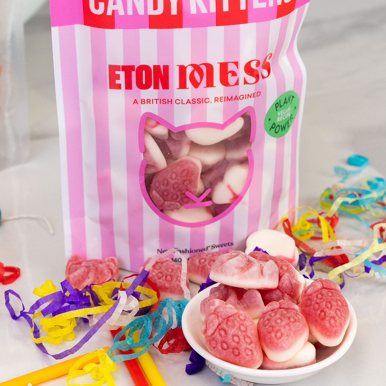 Candy Kittens Eton Mess vegan sweets in a striped pink pouch with strawberry-shaped gummies served in a small bowl alongside colourful confetti.