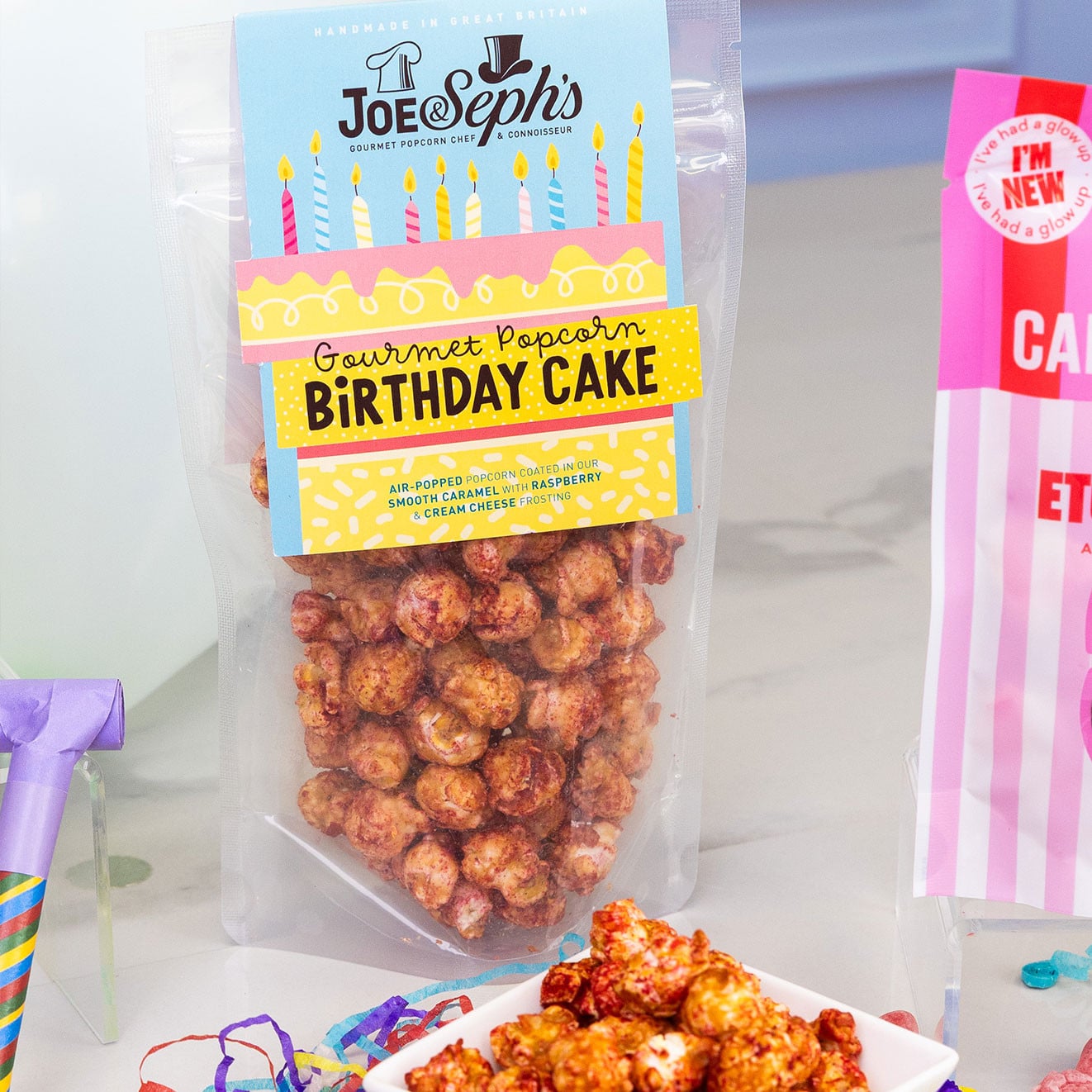 Joe & Seph's Birthday Cake Gourmet Popcorn with raspberry and cream cheese frosting flavour, displayed with a bowl of popcorn and colourful party streamers.