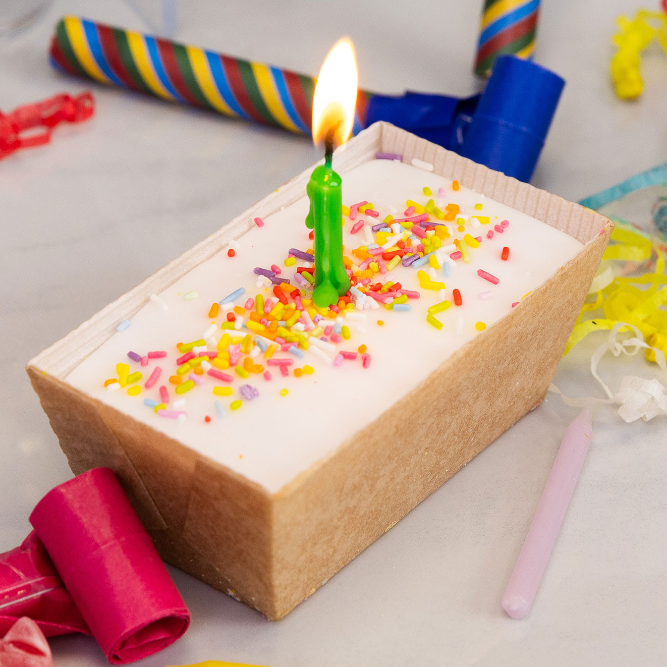 Mini birthday loaf cake topped with white icing, rainbow sprinkles and a lit green candle, surrounded by colourful party blowers and confetti.