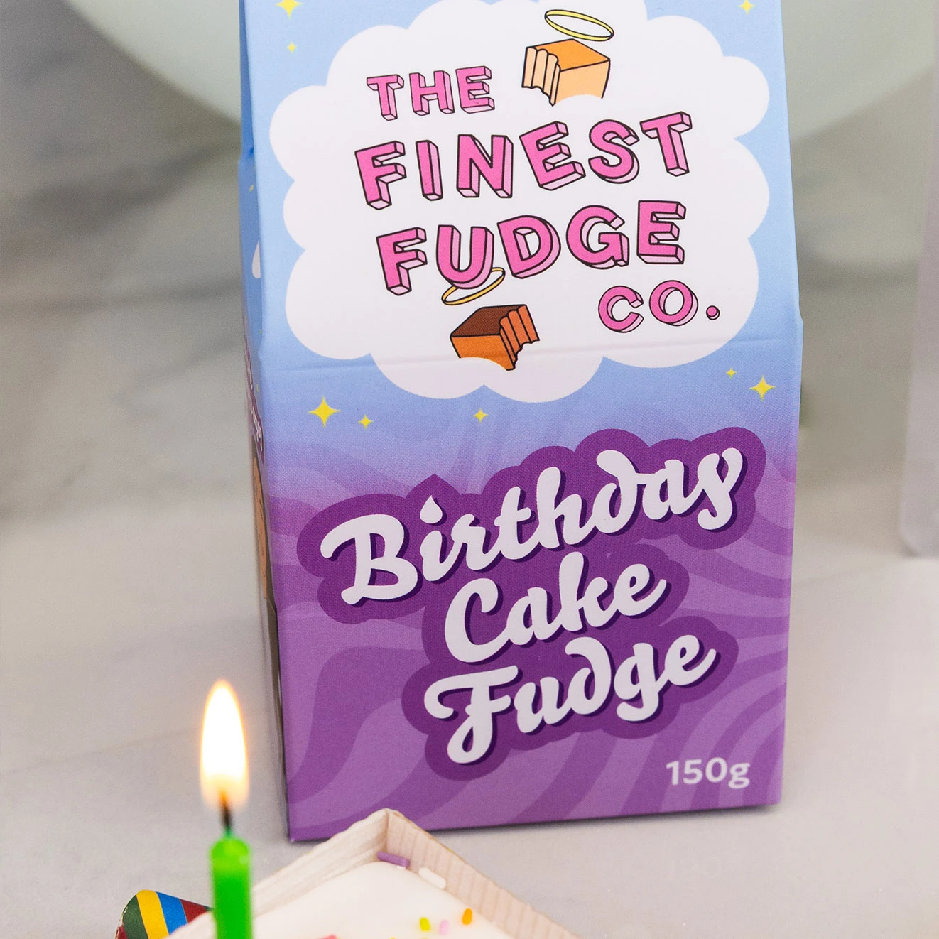 The Finest Fudge Co. Birthday Cake Fudge in a colourful gift box, displayed beside a birthday loaf cake with a lit candle.