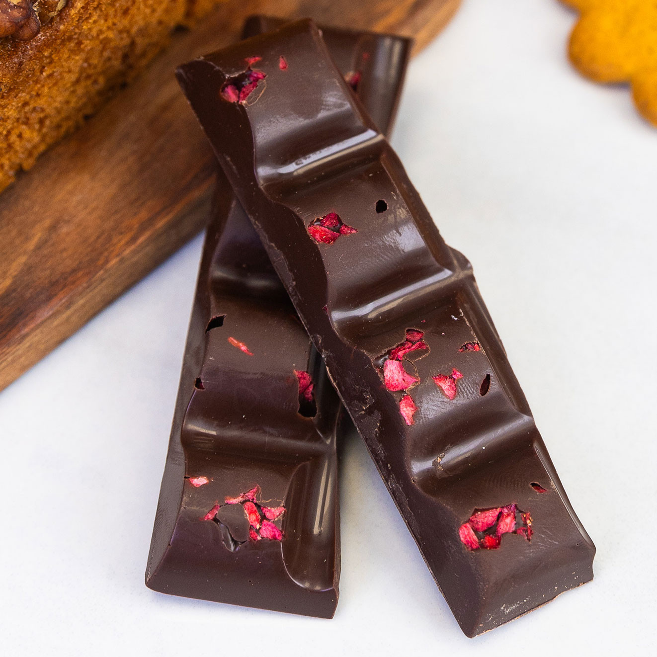 Dark chocolate bars infused with raspberry pieces, stacked beside slices of walnut loaf cake on a wooden serving board.