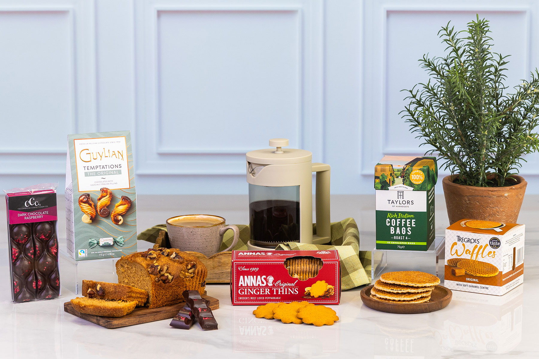 Coffee and treats gift hamper featuring Guylian chocolates, raspberry dark chocolate, walnut loaf cake, ginger thins, stroopwafels, Italian coffee bags, cafetière and a potted plant.